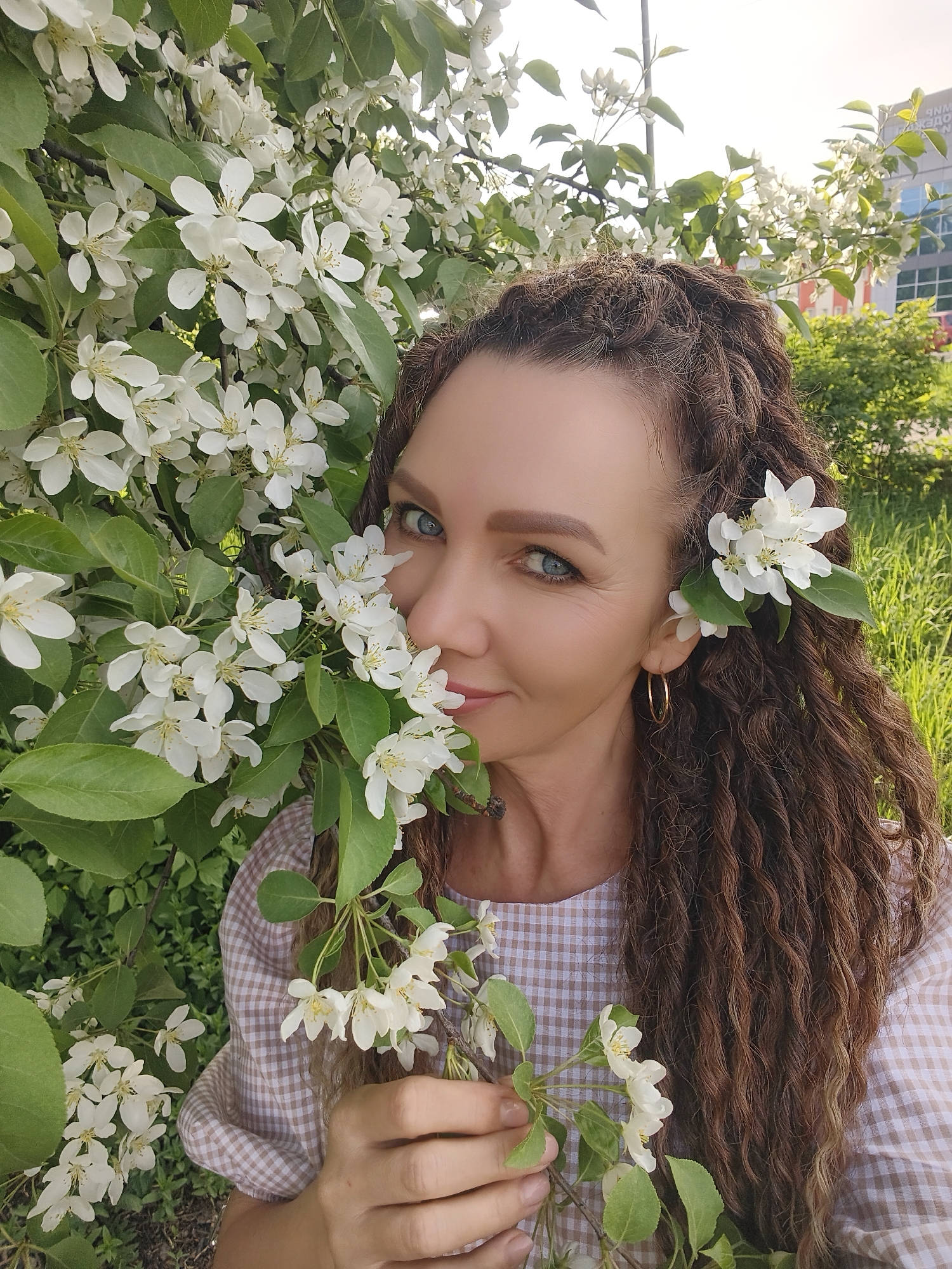 A middle-aged woman with blue eyes and brown, spiral-permed hair stands next to a flowering tree with white blossoms. She holds a sprig of the same blossoms in her left hand. Some blossoms are arranged in her hair near her ears. She wears gold hoop earrings and a light brown and white checkered shirt. The scene is outdoors, with green foliage and a multi-story building in the background, suggesting a city park or garden location.