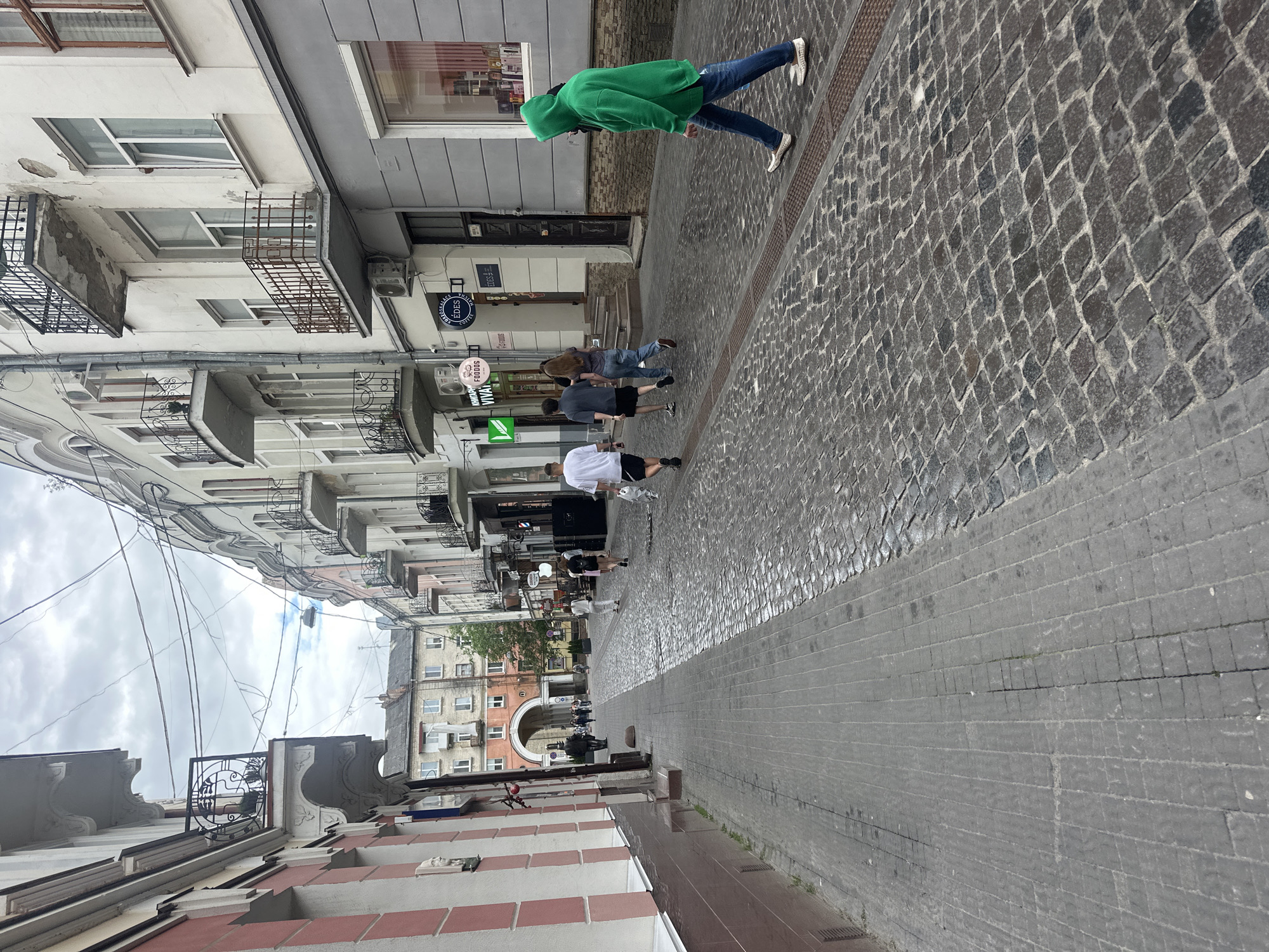 The image depicts a street scene in Ternopil, Ukraine. A narrow cobblestone street is lined with multi-story buildings on both sides. The buildings feature architectural details such as balconies with wrought-iron railings and ornate cornices. Some of the buildings show signs of wear and tear. Several people are walking on the street. In the foreground, on the right side of the street, a person wearing a green hooded sweatshirt and jeans walks away from the camera. To the left, a group of three individuals, two men and a woman, are walking away from the camera. Further down the street, more people are visible, and an arched passageway leads into the distance. The sky is overcast. The street is paved with cobblestones on the right and a different type of paving stone on the left.