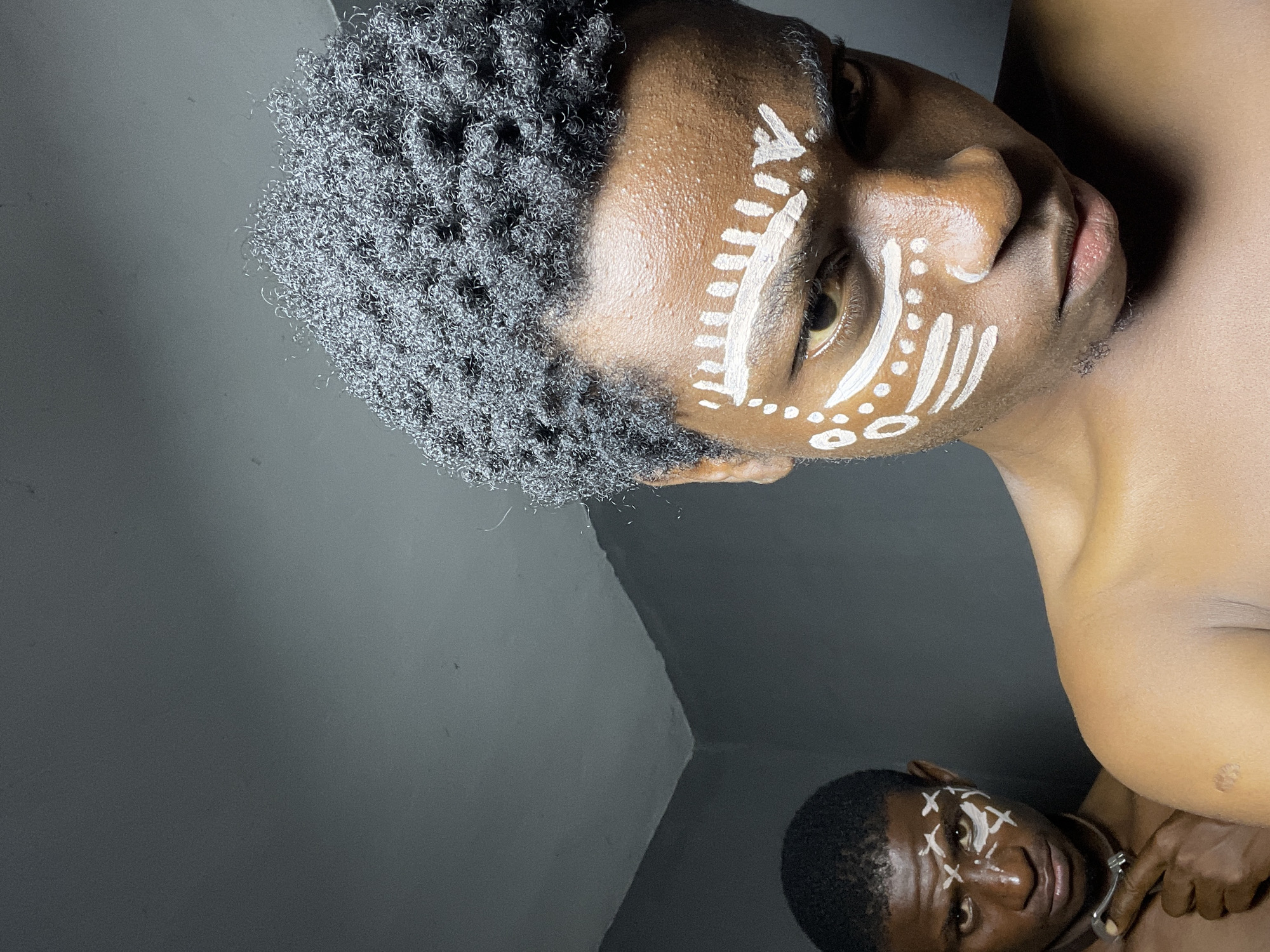 This image depicts two shirtless young men with white face paint markings, in an interior setting that appears to be a corner of a room with gray walls. The man in the foreground, positioned closer to the camera and slightly to the right, has a more detailed white pattern painted on his face, extending from his forehead down to his cheeks and across his nose. He has short, dark, curly hair. The man in the background, to the left, is looking directly at the camera and has a simpler white marking pattern on his face, primarily around his eyes and forehead. He has short, dark, straight hair and is wearing a dark choker-like necklace with a metallic clasp. He is pointing his right index finger towards his left cheek. The lighting suggests a dim environment, with the faces of the subjects illuminated. The location is described as being in Nkpor, Nigeria.