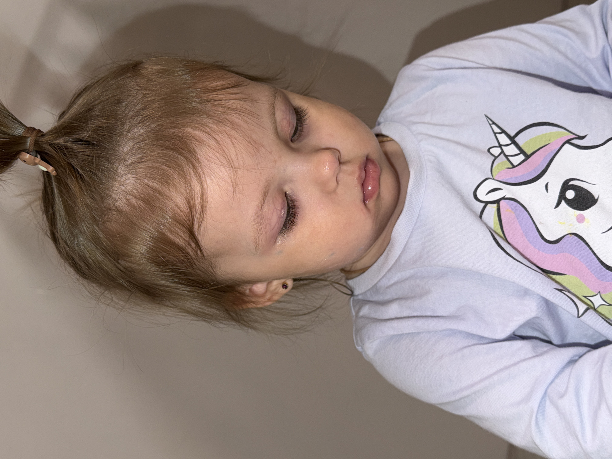 A young child is lying down, eyes closed, wearing a light blue shirt with a unicorn graphic. The child's hair is light brown and pulled back into a small ponytail secured with a brown hair tie. The child has fair skin and a small earring. The background is a plain, light-colored wall. The location is unknown.