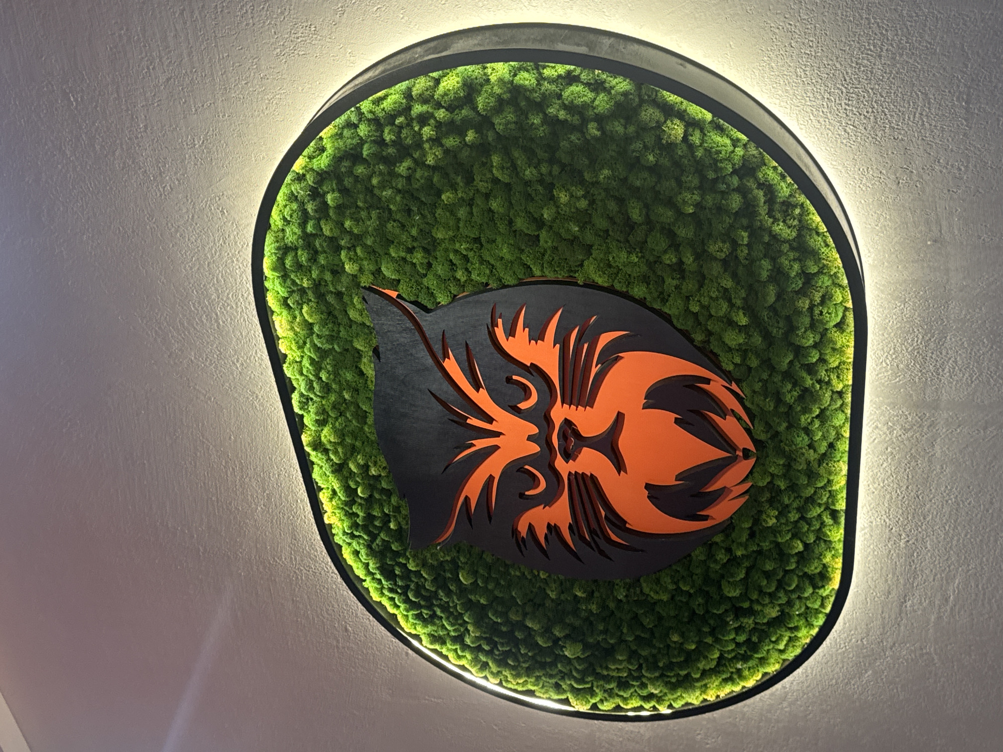 The image shows a decorative wall art piece, possibly indoors in a building in the city of Izhevsk, Russia. The main feature is an irregularly shaped, dark-framed panel filled with green moss. Overlaid on the moss is a stylized, layered logo or emblem of a tiger in black and orange. The wall art appears to be illuminated by a soft light source around the frame. The backdrop is a neutral-colored wall.