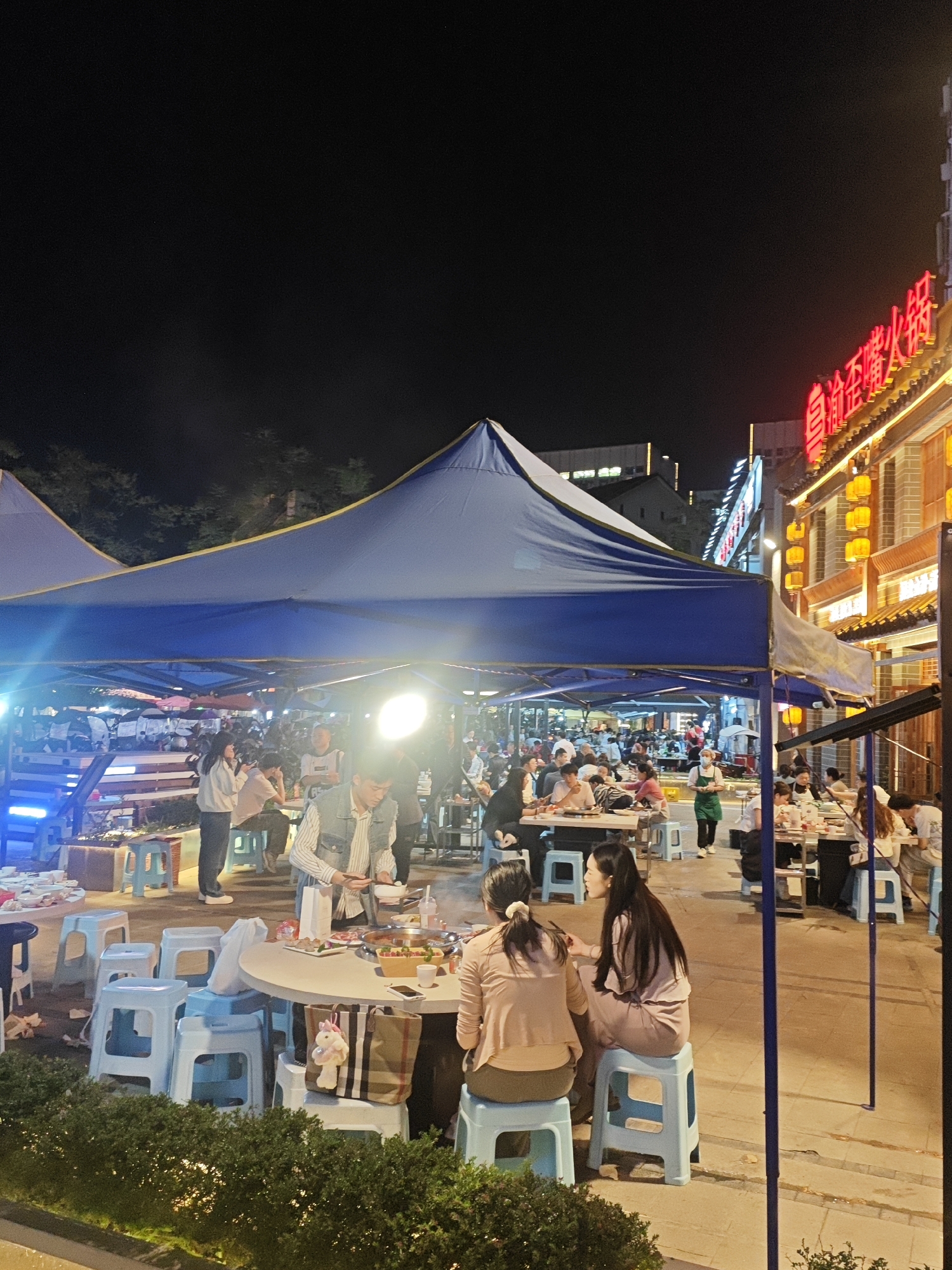 The image depicts an outdoor dining area at night in the Chongqing High-tech Zone, China. Patrons are seated at round tables under blue canopy tents supported by metal poles. Many people are eating. Tables are surrounded by light blue plastic stools. In the background, there is a building with a neon sign in Chinese characters. The environment appears to be a commercial or pedestrian street with trees and other signs of urban life. The scene is illuminated by overhead lights, including a bright light source at the center of the frame.