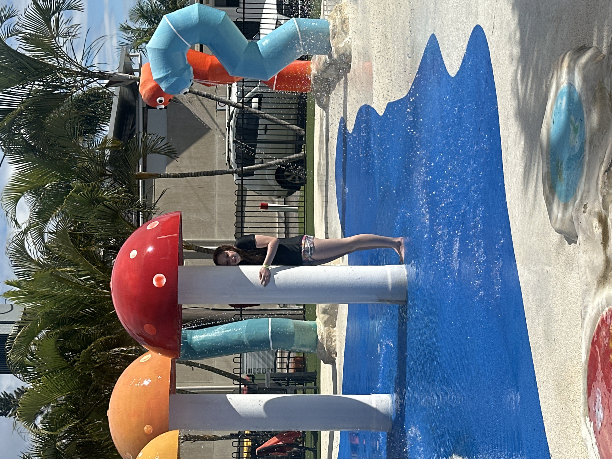 The image captures a section of the Nambucca Heads Main Beach splash pad on a sunny day. The foreground features a bright blue pool of water with white speckles of spray. To the left, a large white mushroom-shaped structure with an orange cap rises from the water, and to its right, a similar mushroom with a red cap is partially obscured by a young woman. The woman, wearing a black t-shirt and patterned shorts, stands with her back to the camera, peeking out from behind the red mushroom. She is holding her arms together in front of her. Behind her, a tall, teal-colored cylindrical water feature with white bands is visible, with water spraying from its top. Further back, an orange inflatable figure with large googly eyes stands next to a blue inflatable figure, both appearing to spray water. A black metal fence runs horizontally across the mid-ground, behind which are palm trees, a building with a gray roof, and parked cars. The ground around the water area is a light-colored, sandy textured surface.