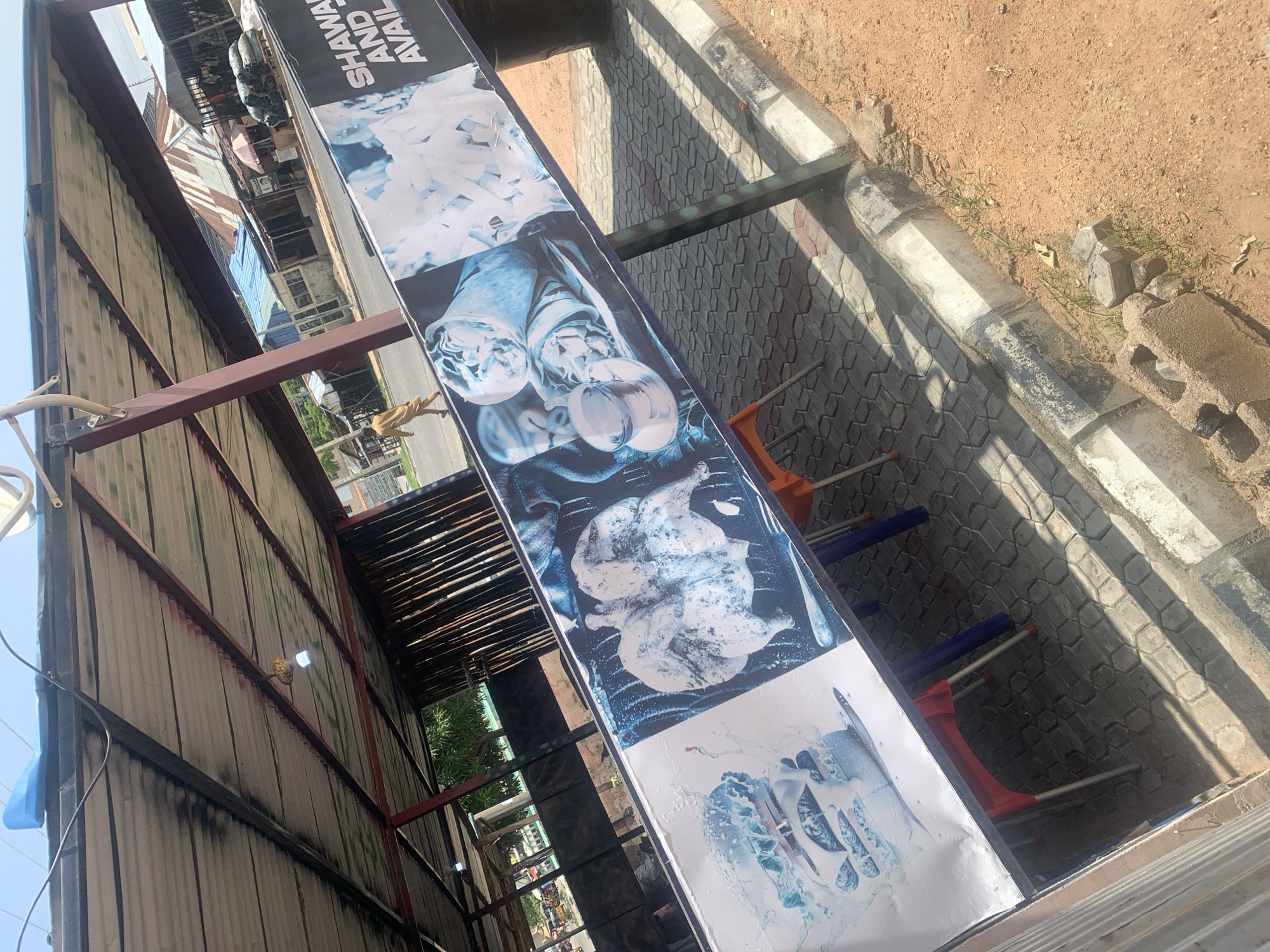 The image shows a street-level view of an outdoor food stall or restaurant in Jalingo, Nigeria. A large banner is displayed diagonally across the foreground, showcasing images of food items like shawarma, burgers, and chicken. The banner appears to be advertising shawarma and other items that are available. Behind the banner, a simple roof structure supported by metal poles is visible, offering shade. The ground is paved with interlocking grey and reddish-brown bricks. In the background, there are several small shops or stalls with corrugated metal roofs, typical of a market or commercial area in a Nigerian city. A person is walking away from the camera on the paved street to the left. The environment appears to be a sunny day, with shadows cast on the ground. The overall scene depicts a typical urban commercial setting in Jalingo, focused on food service.