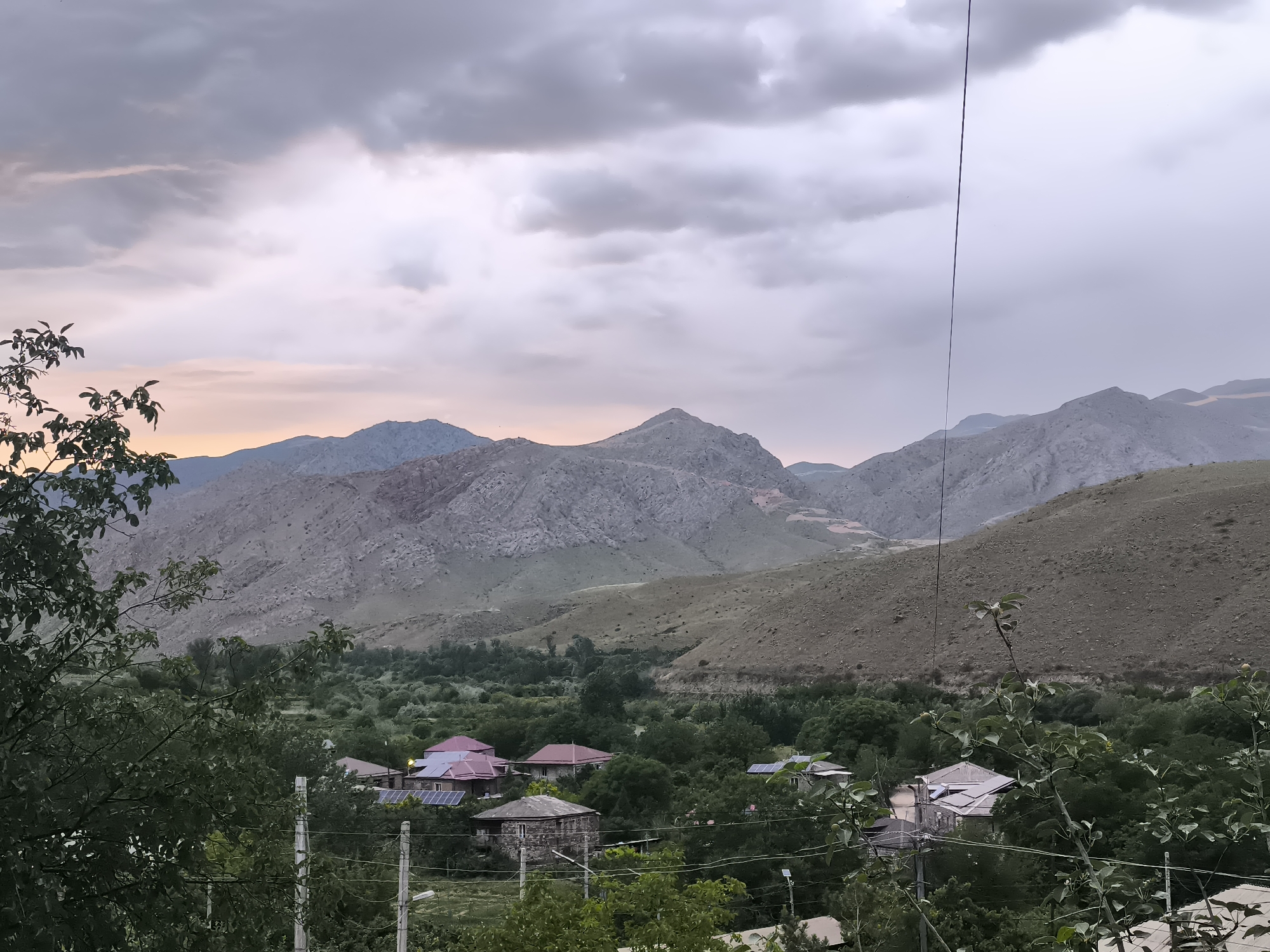 The image presents a scenic landscape of Areni, Armenia, captured during what appears to be late afternoon or early evening under an overcast sky. Heavy grey clouds dominate the upper portion of the frame, with a faint, warm, light purple and orange glow visible near the horizon, suggesting twilight conditions.
In the foreground, dark green tree branches and foliage frame the lower left and bottom sections of the image. Two white posts, possibly part of a fence or gate, are visible on the lower left, and a thin dark utility wire or cable runs vertically down the right side of the frame from the top.
The midground features a verdant valley densely populated with trees, creating a lush green expanse. Several houses are nestled within this foliage, showcasing varied architectural styles and roof colors, including red, light purple, and grey. Notably, solar panels are visible on the roofs of some houses, indicating modern energy adoption in this rural setting. Utility poles and wires are also discernible among the trees.
The background is dominated by a range of large, rugged mountains with rocky, arid surfaces and sparse vegetation, extending across the entire horizon. The mountains appear weathered, with visible contours and geological features. No specific human or animal activity is depicted. The scene conveys a tranquil, settled atmosphere, characterized by the contrast between the green valley and the imposing, rocky mountains under a cloudy sky. No text is visible in the media file.