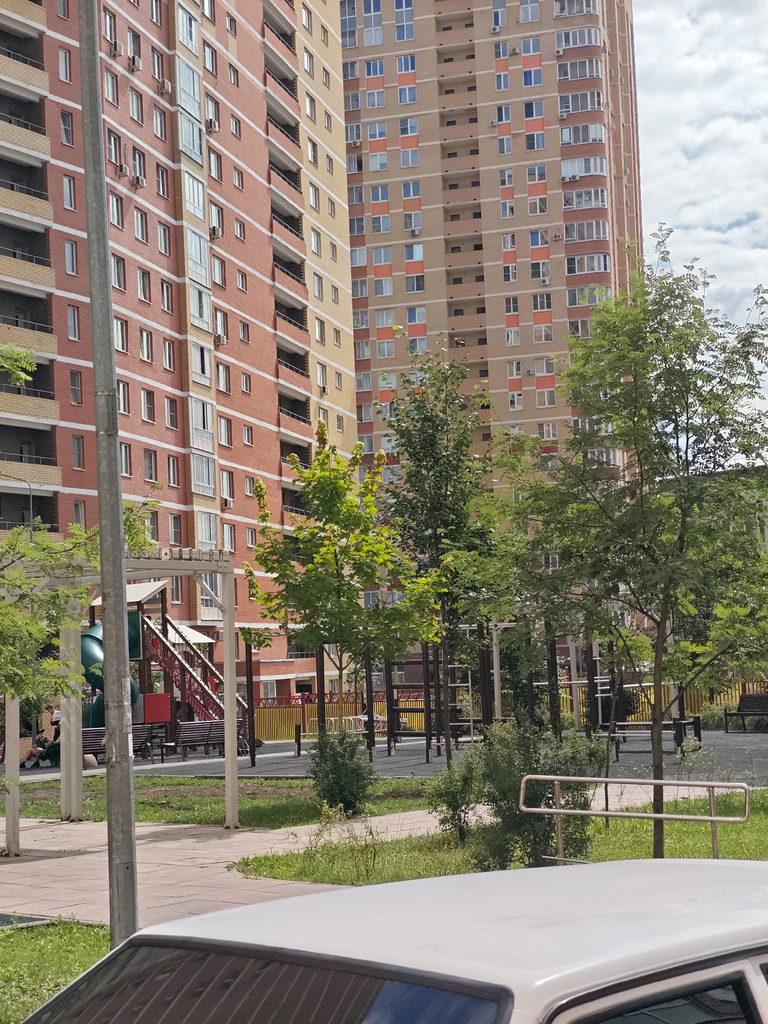 In Shcherbinka, Russia, a daytime scene unfolds in a residential courtyard, featuring two tall apartment buildings constructed with brick and beige paneling. In the foreground, the top of a white car is visible, reflecting the sky. A paved walkway with grassy areas borders the buildings. To the left, a playground with a green slide and red stairs is partially obscured by trees and a light pole. Several individuals are seated on benches near the playground. The background is dominated by the apartment buildings, with a few trees and a fence surrounding the playground area. The sky is partly cloudy.