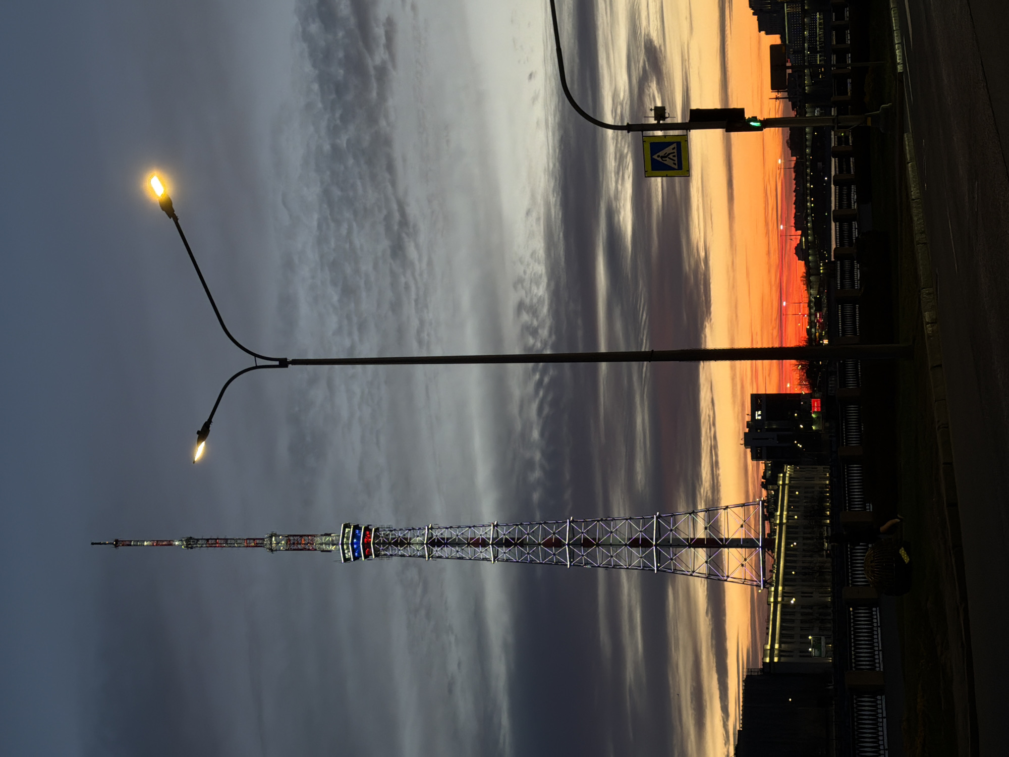 The image depicts a vertical shot of the Saint Petersburg TV Tower against a dramatic sky transitioning from dusk to dawn, featuring a spectrum of dark clouds, orange, and red hues. A Y-shaped street lamp with two lit bulbs is positioned to the left of the tower. A pedestrian crossing sign is visible near the top right of the frame. In the background, there is a cityscape silhouette with buildings lit up against the skyline. The tower itself is a lattice structure illuminated with colorful lights. In the foreground is a dark road or paved area and a decorative fence. The overall scene suggests a city landscape at twilight, capturing the juxtaposition of urban infrastructure with the natural sky.