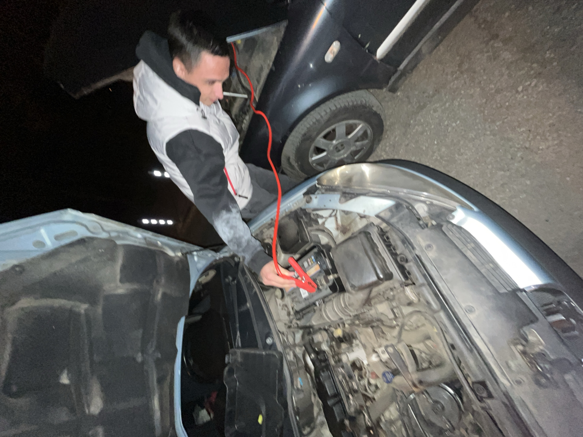 A man is shown connecting jumper cables to a car battery. The hood of the blue car is open, revealing the engine compartment. The man is wearing a light-colored vest over a dark long-sleeved shirt. Another dark-colored car is positioned next to the blue car, with a jumper cable running from its battery. The ground is paved, suggesting an outdoor location. The lighting conditions indicate it is either dusk or night. The location cannot be determined based on the image alone. Sofia, Bulgaria cannot be confirmed based on this image.