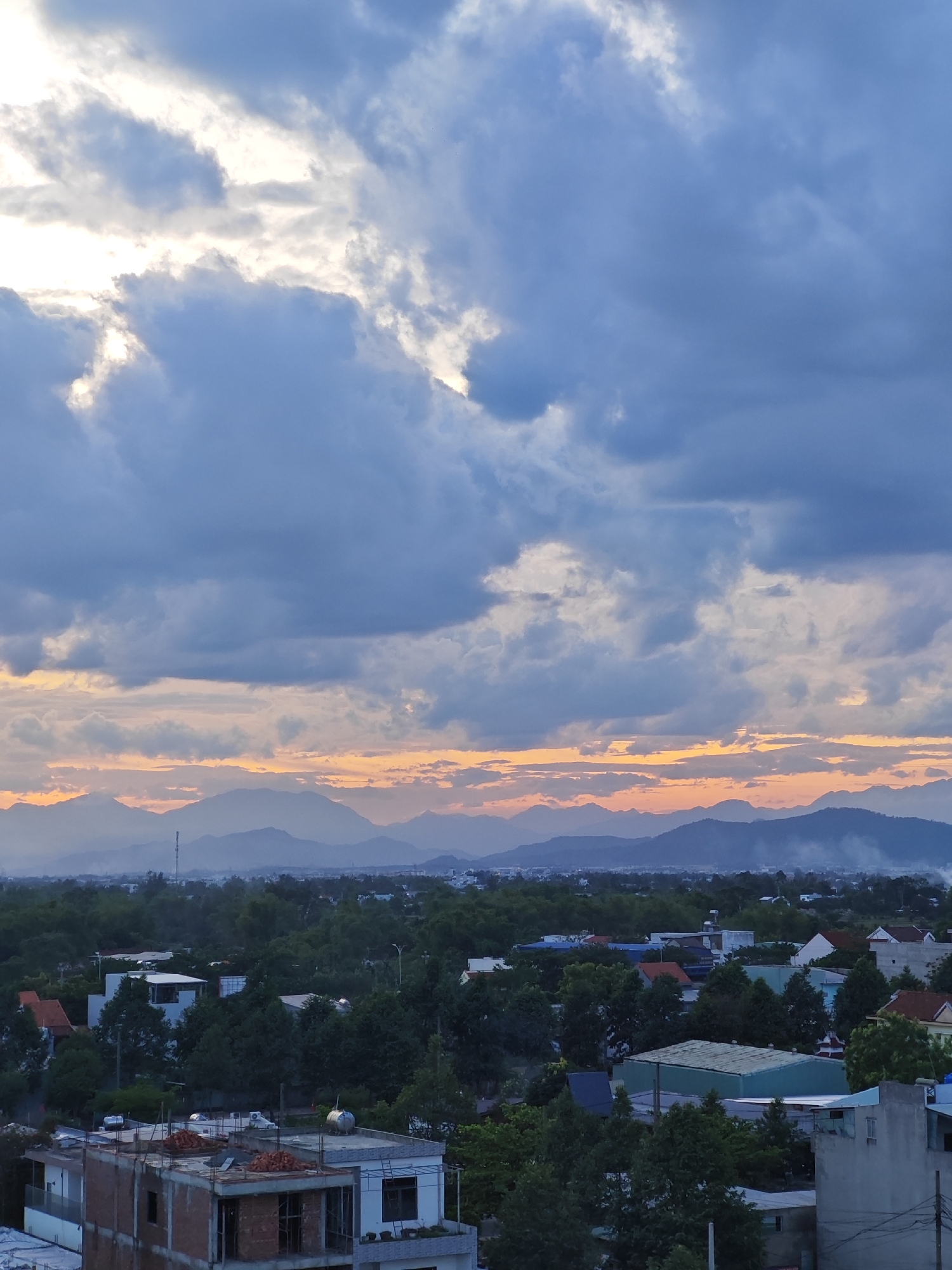 This image depicts an aerial view of a residential area in Da Nang, Vietnam, during what appears to be sunset or sunrise. The sky is filled with dramatic clouds, with patches of bright light suggesting the sun is low on the horizon. The clouds are a mix of dark blues, grays, and whites, with streaks of orange and yellow peeking through, indicative of the twilight colors.
Below the sky, a range of mountains forms a distant backdrop, their peaks softened by atmospheric haze and rendered in shades of blue and gray. The mid-ground is a dense expanse of green trees, indicating a lush, possibly tropical, environment. Scattered amongst the trees are various buildings, mostly residential houses with tiled roofs in shades of orange, red, and white. Some buildings appear to be under construction, with exposed brickwork visible on one foreground structure. A tall communication tower can be seen in the distance among the mountains.
The foreground is dominated by rooftops and the upper floors of buildings. One prominent building in the foreground is under construction, revealing its internal structure and unfinished brick facade. Another building features a white facade and a visible water tank on its roof. The overall impression is of a developing urban or suburban area nestled between natural landscapes. There are no people or animals clearly visible in the image. The scene is quiet and still, capturing a moment of natural beauty during a transitional time of day.