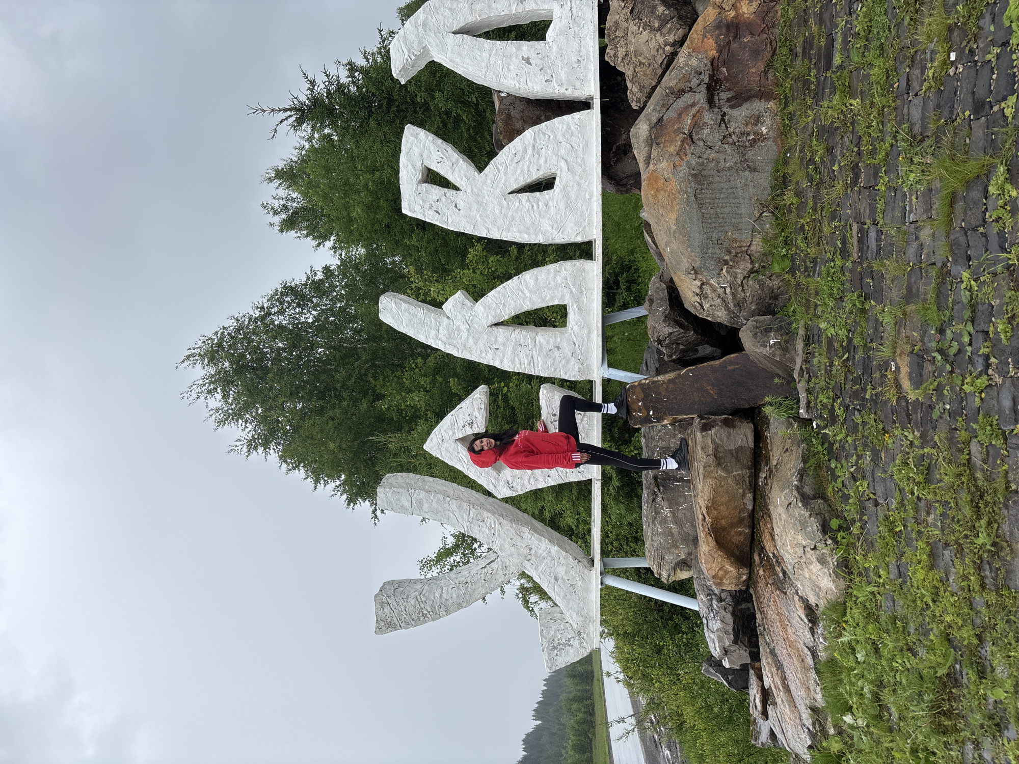 A woman stands on large rocks in front of a white, three-dimensional sign that spells out "ГРЕМЯЧИНСК" in Cyrillic. The sign is made of textured, angular letters, with the woman positioned in front of the first letter. She is wearing a red hooded jacket, black leggings with white stripes, and black sneakers with white socks. Her left foot is placed on a large rock, and her right leg is bent. The sign is supported by a white metal frame that rests on a collection of large boulders. Behind the sign, a dense forest of green trees covers a hill. The sky is overcast and gray. To the left of the sign, a paved road is visible, bordered by more greenery. The foreground shows dark, rough paving stones interspersed with patches of green grass. The overall scene suggests an outdoor location in a natural, possibly rural or mountainous area, marked by the prominent town sign.