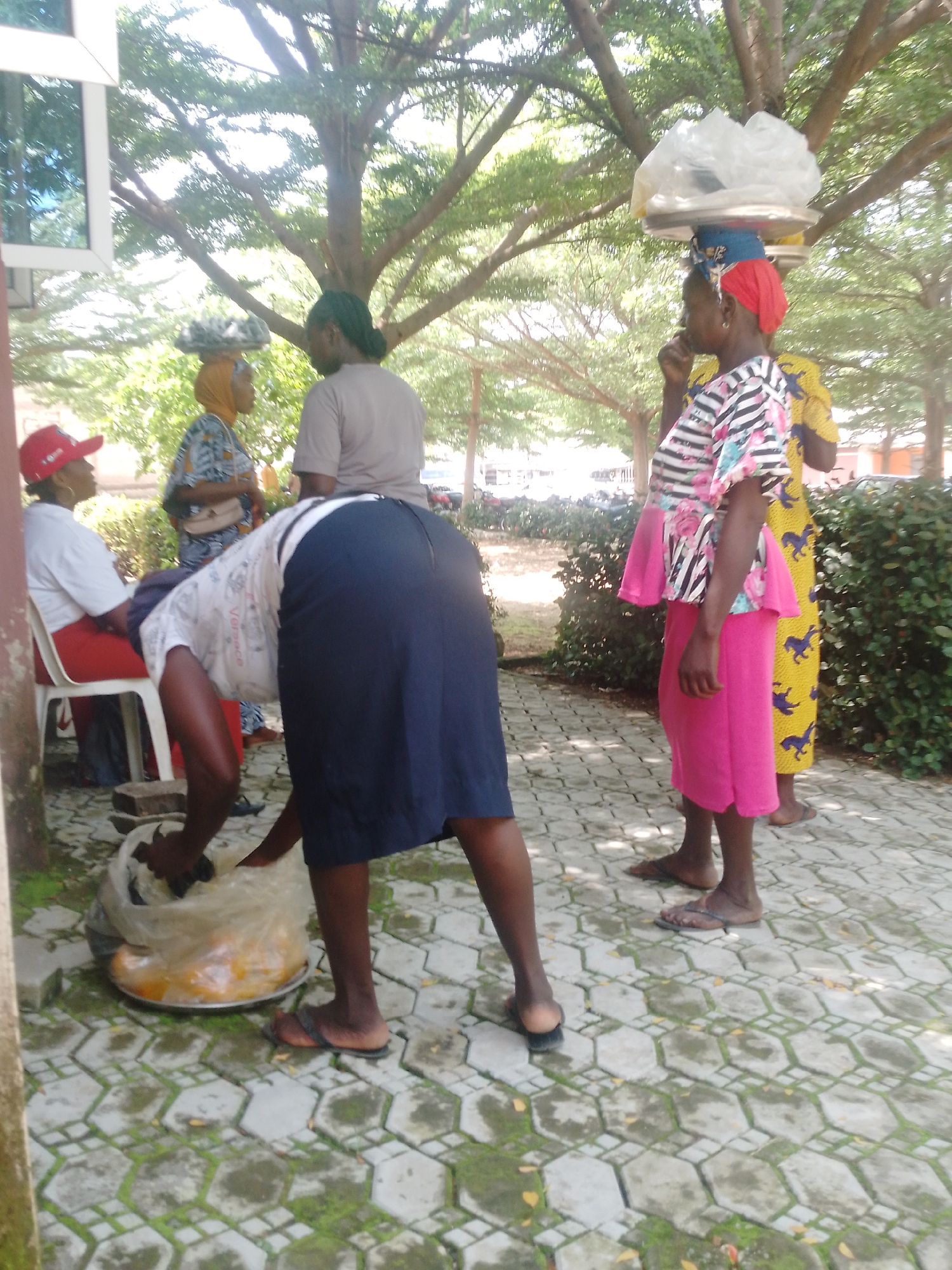 In Akwanga, Nigeria, four women are gathered outdoors. One woman, wearing a red cap and white shirt, sits on a red and white chair. Another woman, in a blue and white patterned dress and yellow headscarf, stands nearby. A third woman, dressed in a light brown shirt and green headscarf, stands to the left of the center. The fourth woman, wearing a dark blue skirt and a white shirt with colorful patterns, is bent over attending to a large tray of yellow produce covered in plastic. Two women are seen from the side with headscarves and clothing that suggests they are vendors, one with a red headscarf and a patterned shirt, the other in a yellow patterned dress. The ground is paved with hexagonal tiles with moss growing in the gaps. Trees with large branches and green foliage provide shade. The scene appears to be a marketplace or roadside vending area.