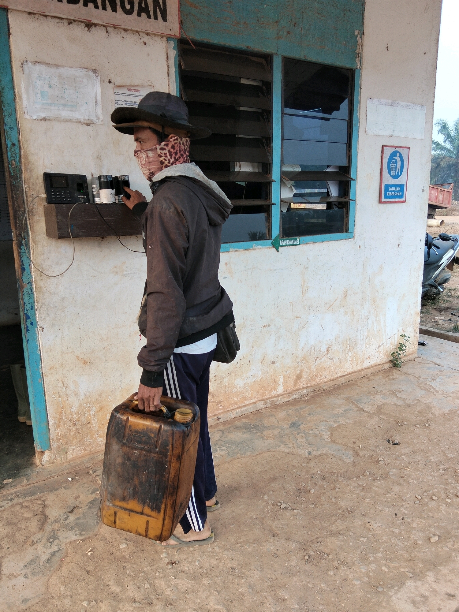 In Teluk Tenggirik, Indonesia, a man stands near a building with white walls and a blue-trimmed window. He wears a dark jacket, dark pants with white stripes, a dark hat, and a leopard-print face covering. He holds a dark yellow, old plastic jerrycan in his right hand and is reaching for a black device mounted on a wooden plank attached to the wall. This device appears to be a time clock or similar electronic equipment. Behind him, the building has a dark blue doorframe on the left and a blue-trimmed window with louvered panes. A blue and white sign with a trash can icon and text in Indonesian is visible on the wall to the right of the window. The ground is a mix of concrete and dirt. A motorcycle is parked to the far right. The overall environment suggests an outdoor, possibly semi-rural or industrial, setting.