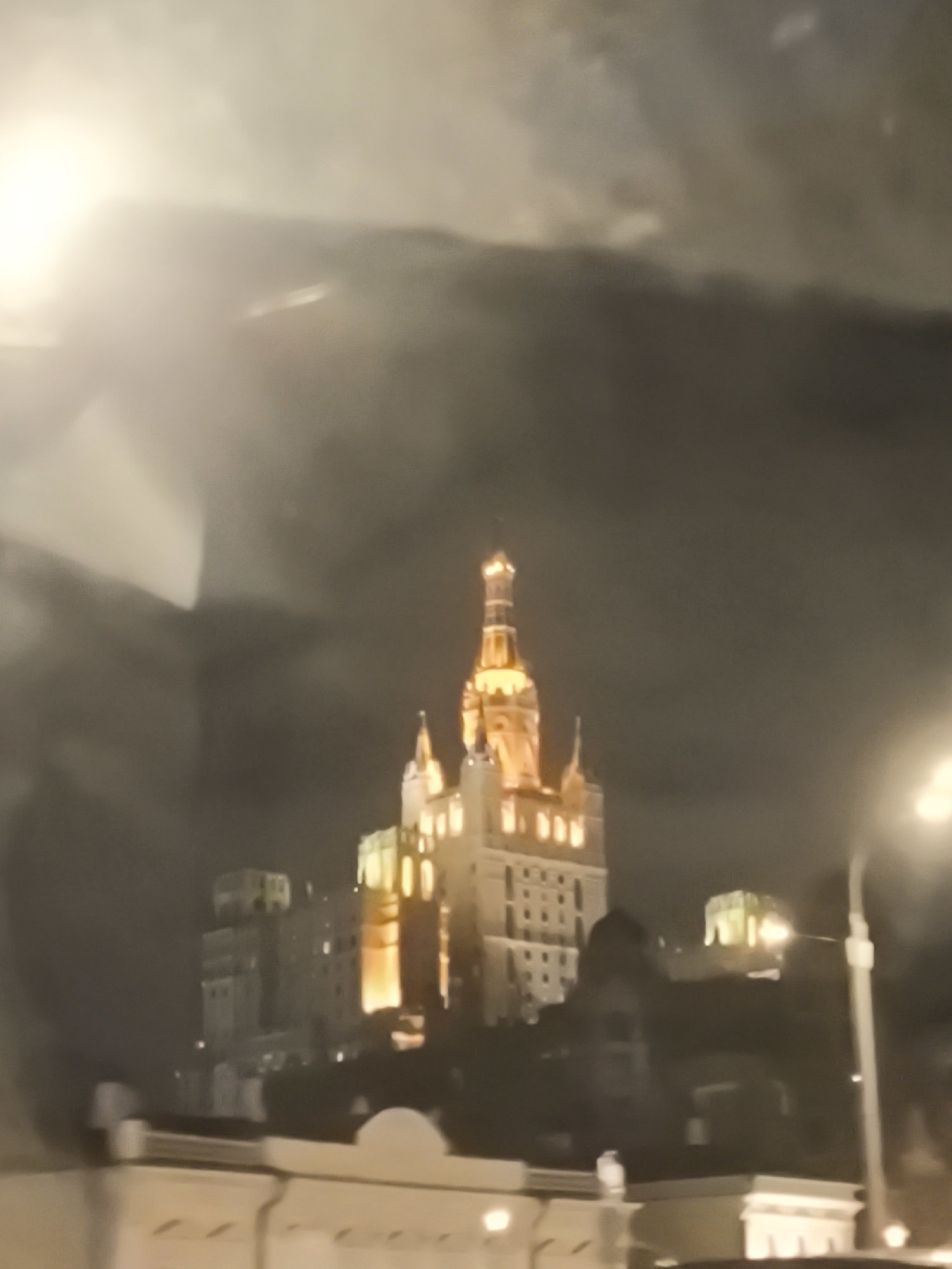 This night-time image captures a grand, illuminated building, characteristic of Moscow's Stalinist Empire style, against a dark, cloudy sky. The central structure, strongly resembling one of the city's iconic "Seven Sisters" skyscrapers (likely the Hotel Ukraina, also known as Radisson Collection Hotel, Moscow), dominates the frame with its imposing height and elaborate architecture. Its multi-tiered spire and intricate facades are bathed in warm, golden light, creating a striking contrast with the obscured, somber clouds above.
Smaller, less distinctly lit buildings are visible in the background and to the sides, indicating a dense urban environment. In the blurred foreground, light-colored architectural elements and a street light on the right edge suggest the vantage point is from a street level or a nearby structure.
The photograph is marked by significant light reflections and general haziness, particularly in the upper left and right, implying it was taken through glass, possibly a window or a vehicle's windshield. There are no people visible, and no specific activity is taking place beyond the static illuminated scene. The overall impression is one of architectural majesty and historical significance, set against the dramatic backdrop of a cloudy Moscow night, reinforcing the city's unique architectural heritage. No visible text is present.