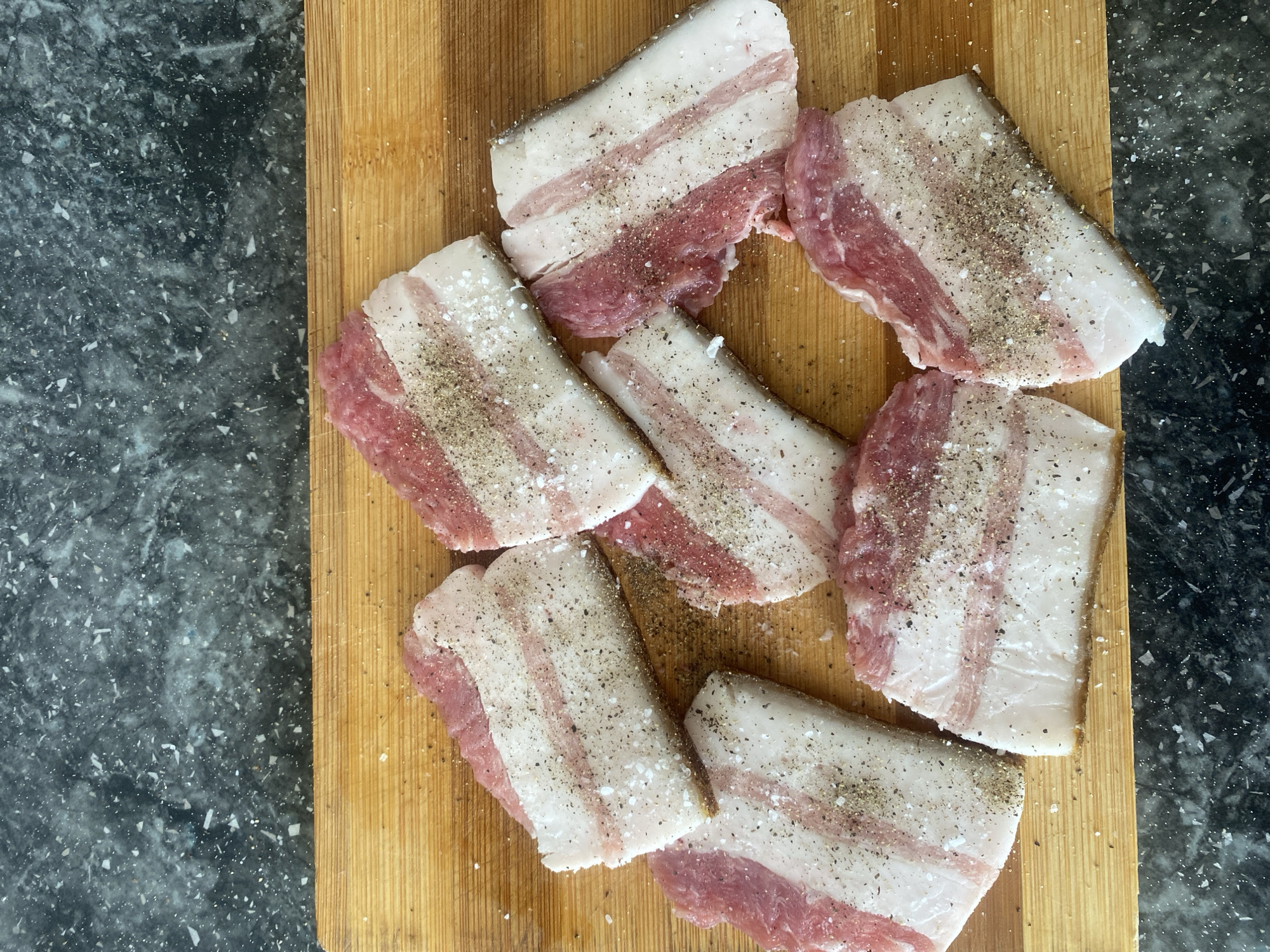 The image shows several slices of raw bacon seasoned with salt and pepper. The bacon is arranged on a wooden cutting board, which is placed on a dark, speckled countertop. The bacon slices have distinct layers of white fat and pink meat. The seasoning is visible as white salt crystals and dark pepper specks across the surface of the meat. The lighting appears to be overhead, casting subtle shadows. The scene is a close-up, overhead shot, focusing on the food preparation. There is no visible text in the image. The overall impression is one of simple food preparation, possibly for cooking.