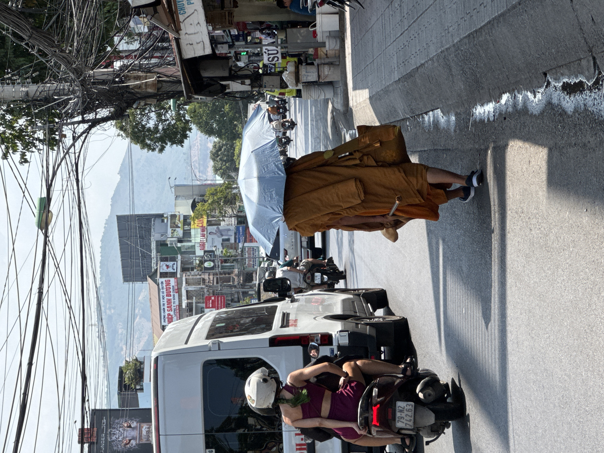 The image captures a street scene in Nha Trang, Vietnam, under a bright sky, with a focus on the bustling traffic and pedestrian activity. In the foreground, a person wearing a saffron-colored robe and carrying a bag walks away from the camera, holding a light blue umbrella overhead. They are on a sidewalk to the right of a street.
On the street, a white van is visible on the left, and a woman on a scooter wearing a helmet and a dark red outfit is seated on it, facing away from the camera. Other motorcycles and a car are also in the traffic. The background shows buildings, advertisements, and a hilly landscape. A dense network of overhead electrical wires crisscrosses the scene. The road surface is asphalt, with a concrete sidewalk and a concrete curb separating it from the street.
