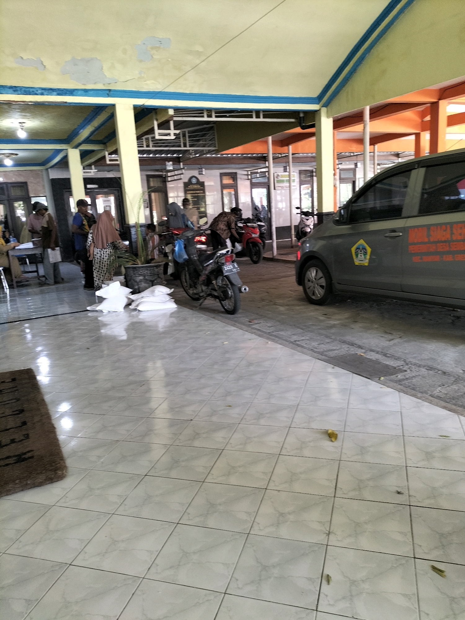 This is a photograph taken at a building in Sembayat, Indonesia. The scene appears to be an open-air pavilion or covered walkway adjacent to a building. The floor is tiled with a light-colored marble pattern. In the foreground, a dark brown doormat is visible.
Several people are present in the scene. To the left, a person wearing a pink shirt and dark pants stands near a desk where another person is seated. Further back, a person in a blue shirt and black pants is visible. A woman wearing a patterned dress and a headscarf stands near a potted plant. Other individuals are seen in the background, some near motorcycles.
Several motorcycles are parked in the area. One motorcycle is in the center of the frame, and another is further to the left. A gray car is parked on the right side of the image. The car has text and a logo on its side. White bags, possibly filled with rice or other goods, are stacked on the tiled floor.
The architecture includes yellow painted pillars and beams, with blue trim along the ceiling edges. The ceiling itself is painted yellow and shows signs of peeling paint. In the background, part of the building features orange architectural elements. Lighting fixtures are visible on the ceiling. The overall impression is that of a public or community building, possibly a sub-district office or a similar administrative center.