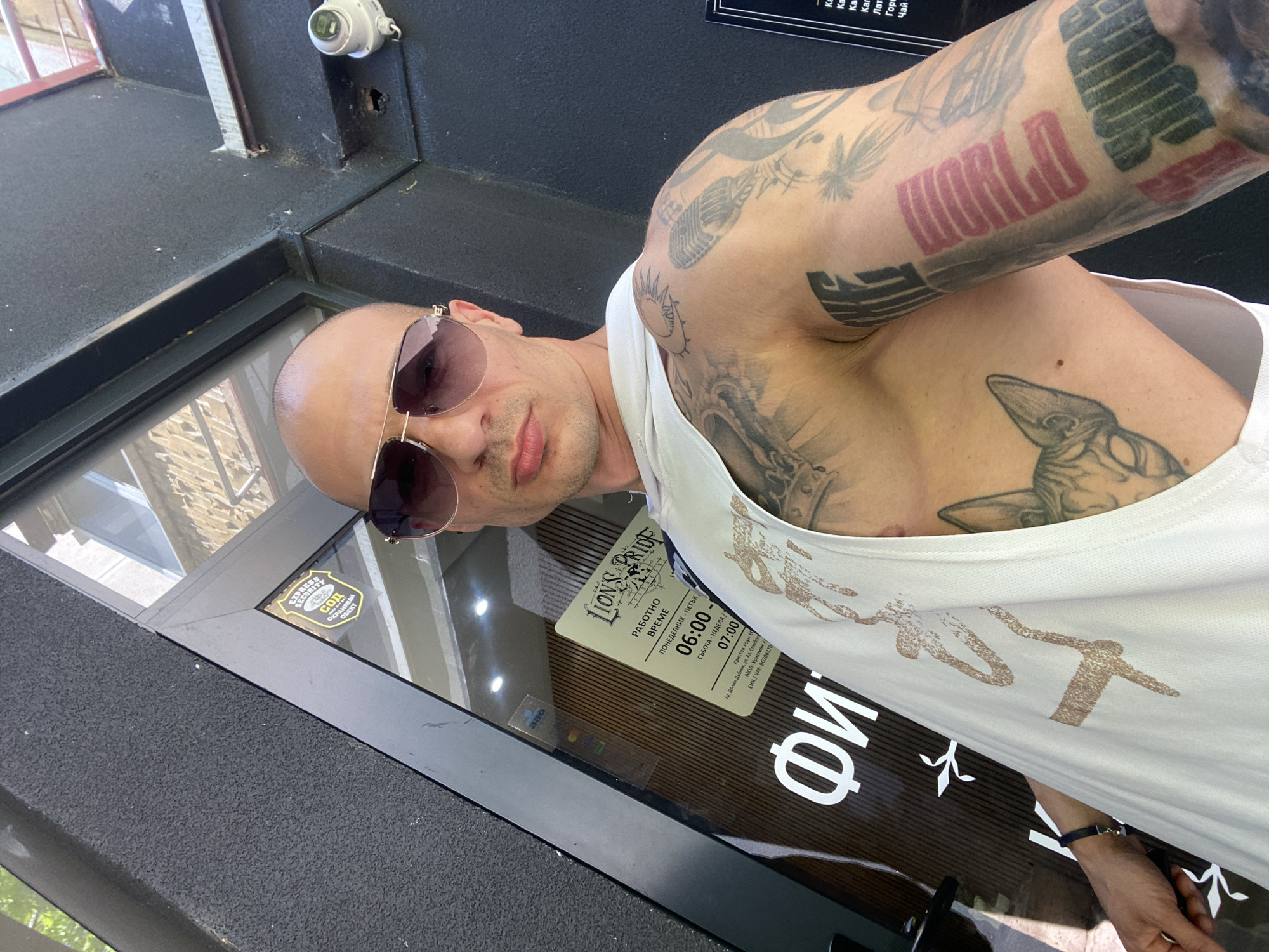 A bald man wearing sunglasses and a white tank top with a partial chest and arm tattoo is posing in front of a storefront. The storefront has a sticker for "EXPRESS SECURITY СОД" and business hours for "LION'S PRIOF", a company located at "Гр. Долни Дъбник, ул. Ал. Стамболь" and likely involved in security services. The street name may correspond to Aleksandar Stamboliyski street in Dolni Dabnik, Pleven Province, Bulgaria. A security camera is mounted above the storefront.