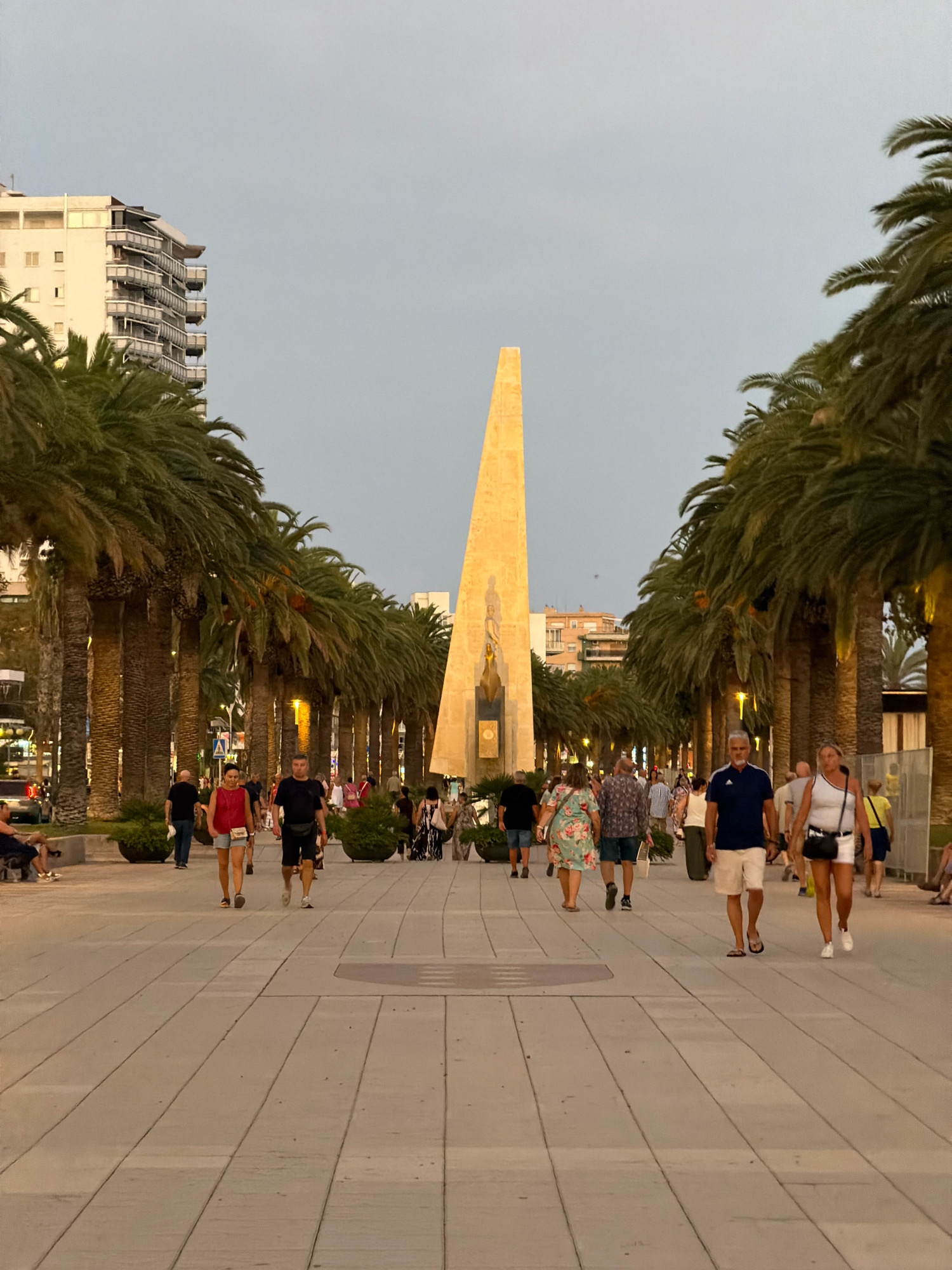 A vertical, full shot captures a wide, pedestrianized promenade lined with tall palm trees on either side. In the center of the promenade, an illuminated, tall, golden obelisk-like monument stands prominently, featuring a statue at its base. People are walking in both directions along the promenade, dressed in casual summer attire. The scene is set during dusk, with a soft, hazy sky and artificial lights illuminating the path and monument. In the background, a multi-story building with balconies is visible, as are other structures and a distant vehicle. The promenade is paved with light-colored rectangular tiles. The environment suggests a popular tourist area in Salou, Spain, likely a waterfront promenade, given the palm trees and open public space.