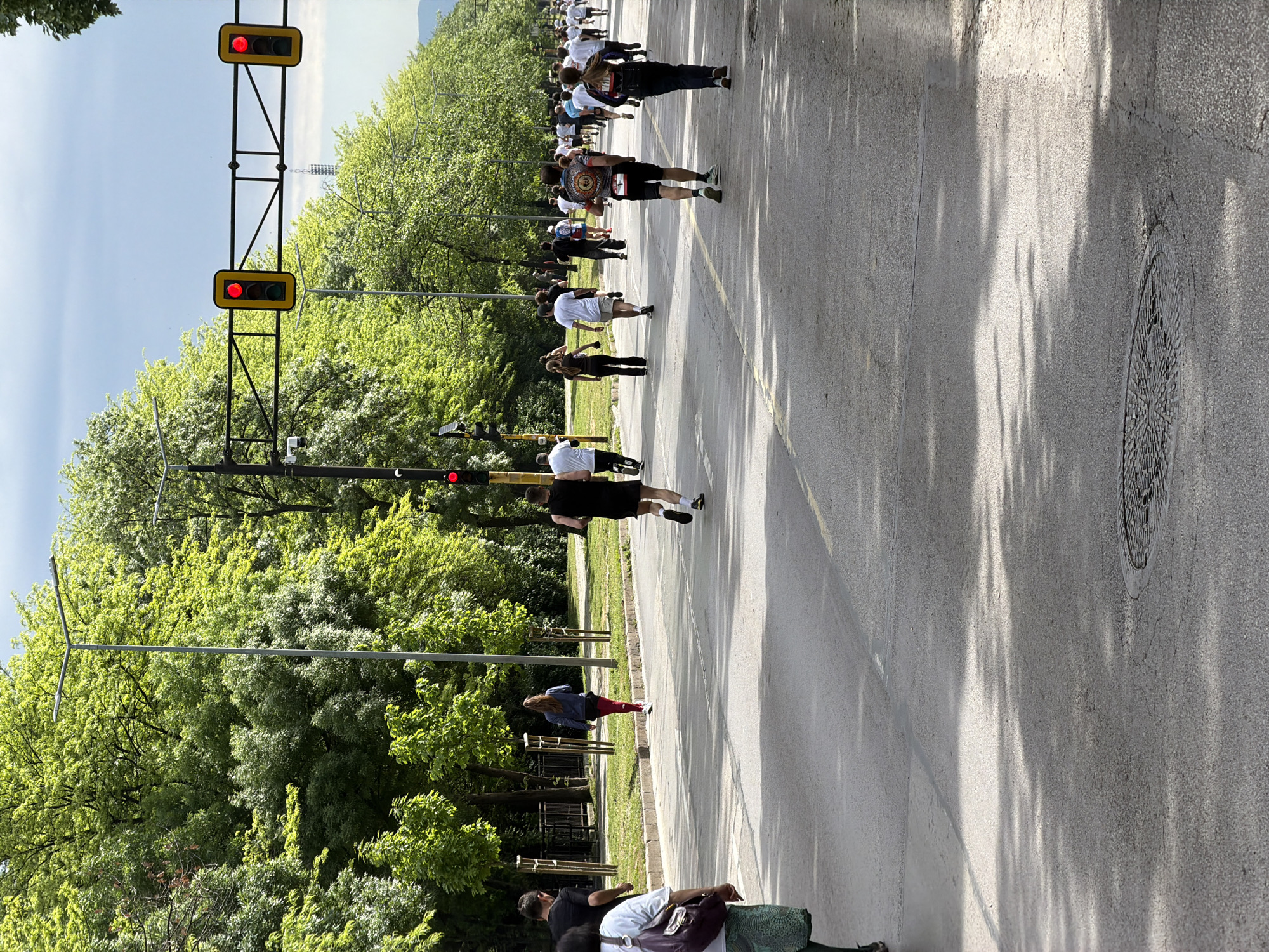 The image shows a street in Sofia, Bulgaria, where a large group of people are participating in a running event. The runners are moving away from the camera. The street is lined with trees on the left side. A traffic light stands on the left with both signals showing red. The sky is visible at the top of the image, appearing bright and clear. A manhole cover is visible on the right side of the street. In the bottom of the frame there are people standing on the left side of the street with some personal belongings.