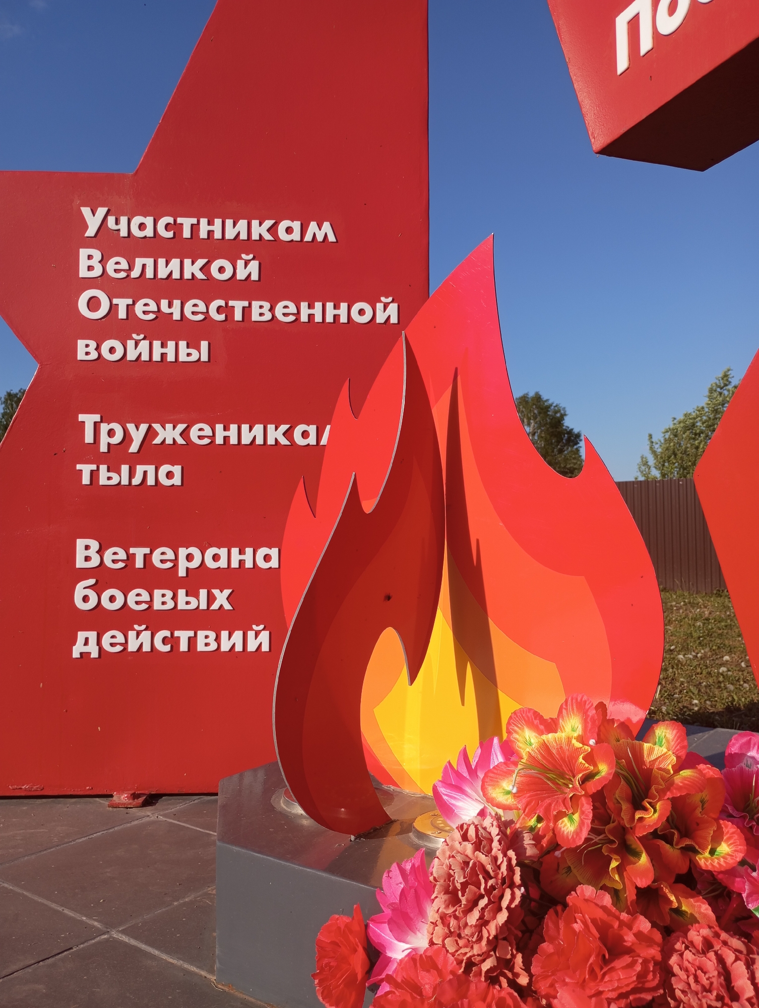 A photograph depicts a monument in Буренка, Russia. The monument is composed of a red star-shaped sign with white Cyrillic lettering that translates to "Participants of the Great Patriotic War," "Workers of the Rear," and "Veterans of Hostilities." Next to the sign is a representation of a flame made of layered red and yellow material, and an arrangement of red and pink artificial flowers lies at the base of the flame. The background shows a blue sky, trees, and a fence. The monument stands on a tiled platform.