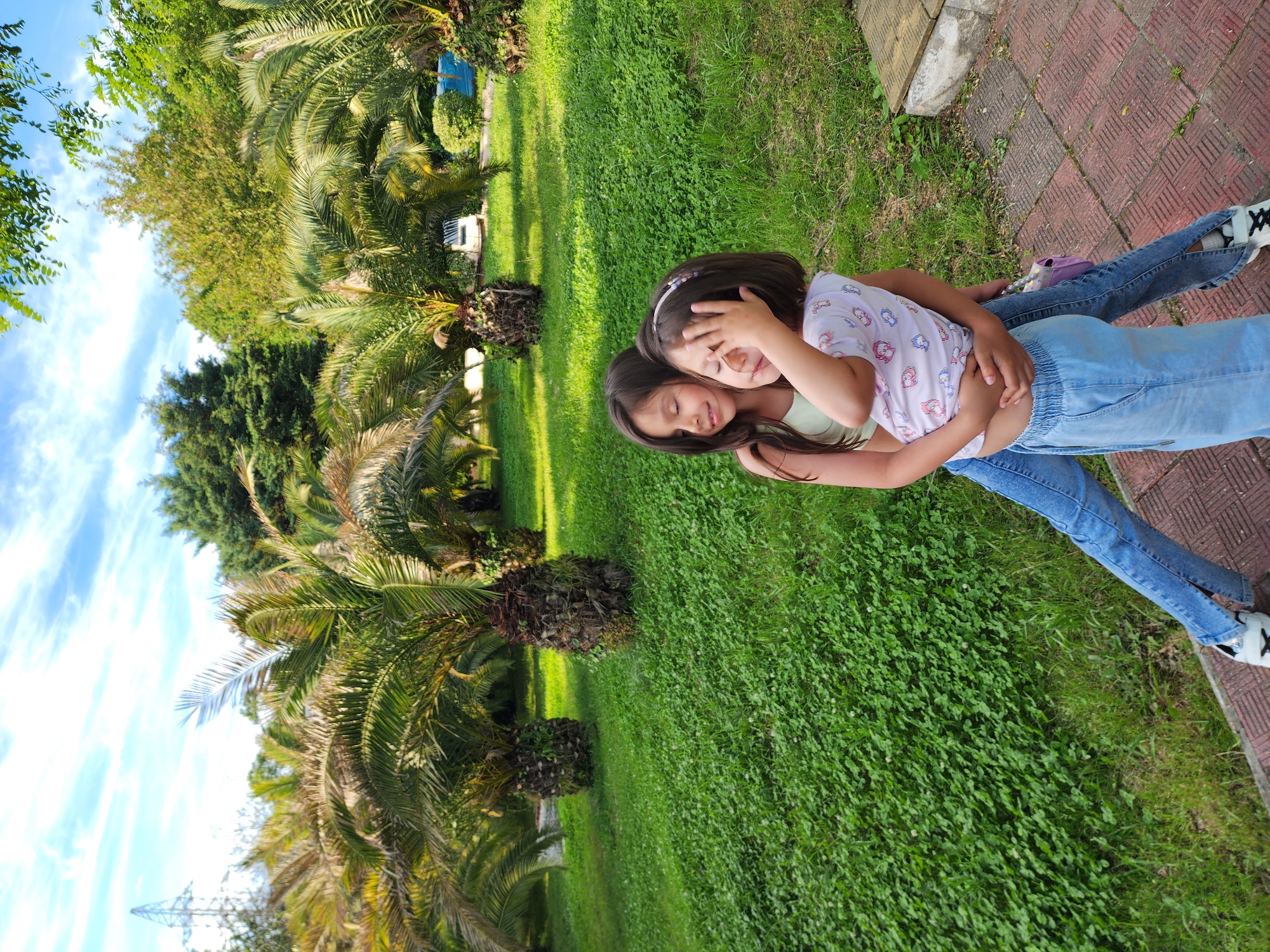 The image captures two young girls embracing each other in a vibrant outdoor park or garden under a clear blue sky with scattered white clouds, suggesting a sunny day.
The girl in the foreground, wearing a light green top and blue jeans with white sneakers, is being hugged from behind. She has dark brown hair and her left hand is playfully covering her face, with her eyes closed or partially closed, conveying a sense of shyness, affection, or joy. The other girl, who is doing the hugging, has dark brown hair with a light-colored headband and wears a light-colored t-shirt with various cartoon-like patterns. Her head is resting gently on the first girl's shoulder, and her arms are wrapped around her waist.
The setting is characterized by lush green grass that fills a significant portion of the frame, along with numerous palm trees and other dense green foliage in the background and mid-ground. A paved pathway made of red brick-like stones is visible along the right edge of the image. In the distant background, a metallic structure, possibly a power tower, can be faintly seen, along with some distant structures like a blue object and a white block. The presence of numerous palm trees aligns with the subtropical climate found in Sochi, Russia. The overall mood of the image is warm, affectionate, and playful. There is no clearly visible text in the image.