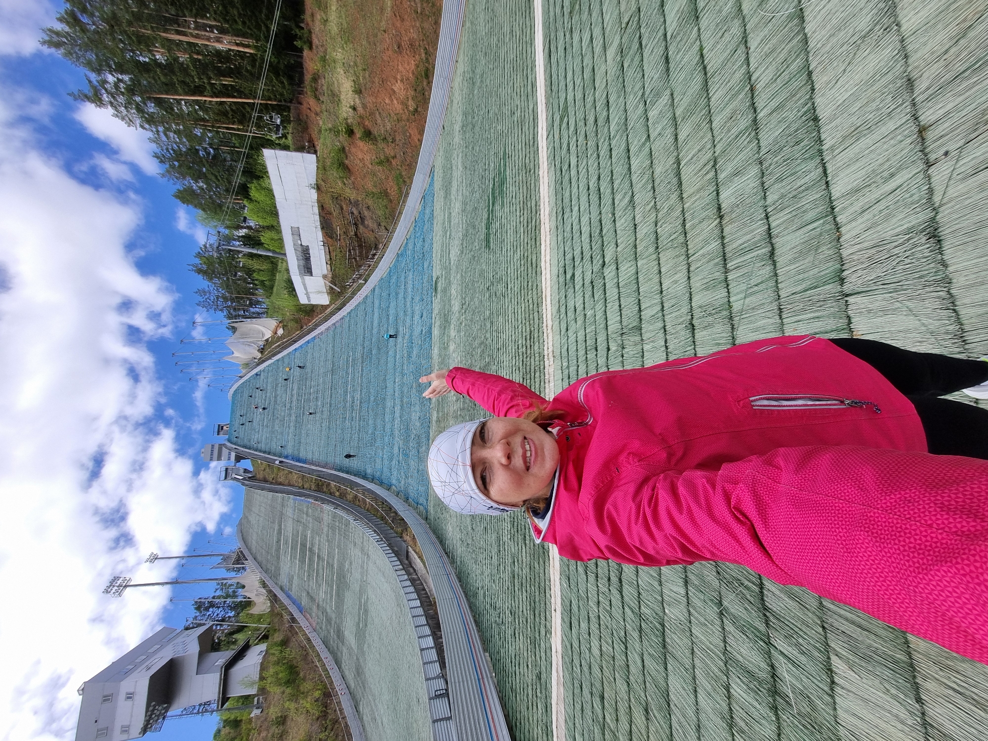 A woman in a pink jacket and a white patterned cap is standing on the grass surface of a ski jump in Прикамский, Russia. She is pointing towards the top of the ski jump. The ski jump's grassy surface has lines running down its length. In the background, there is a cloudy sky, tall trees, and buildings associated with the ski jump facility.
