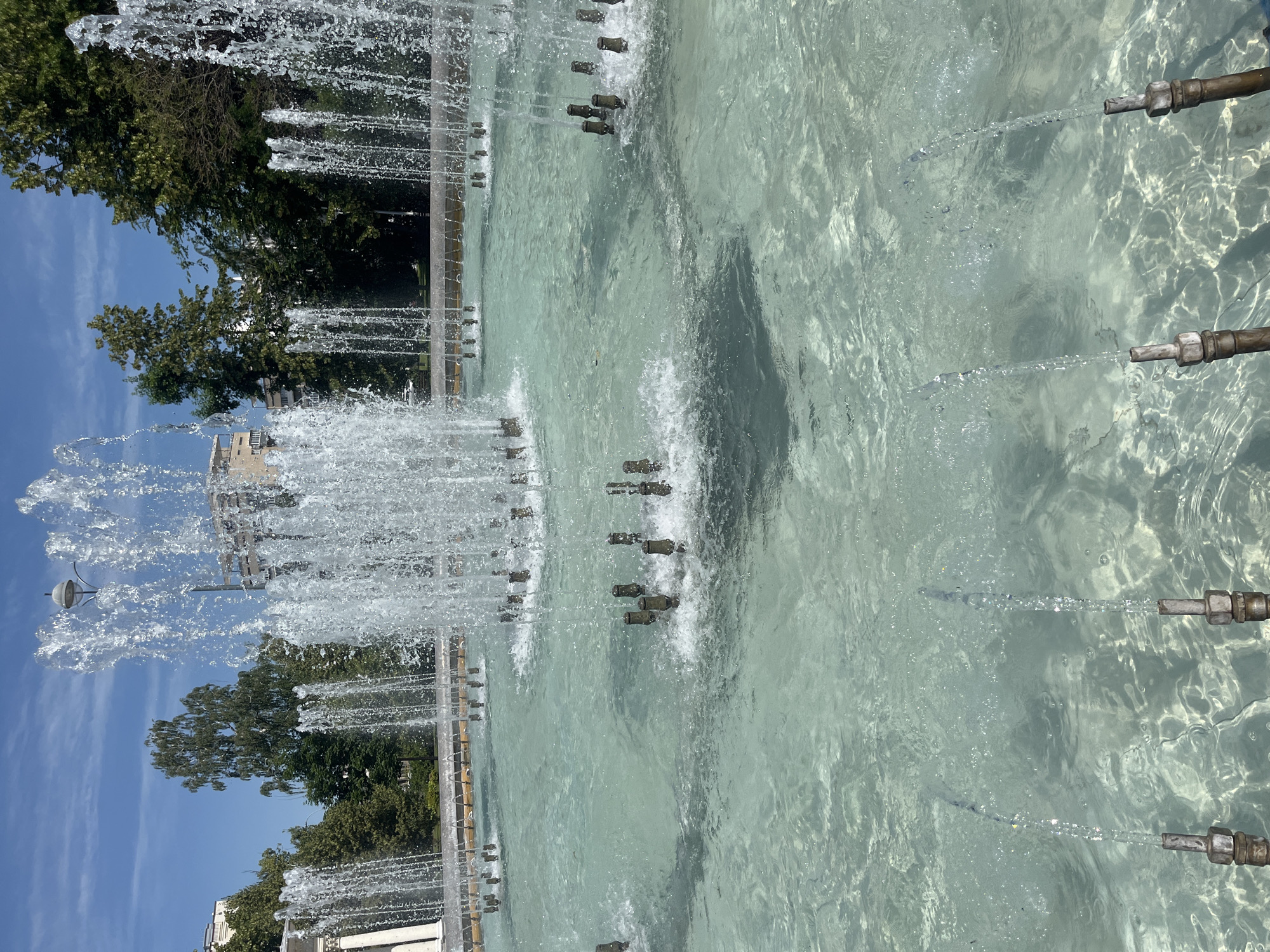 The image shows a public fountain with multiple jets of water spraying upwards in a large pool of clear, turquoise water. The fountain is surrounded by trees and buildings, suggesting an urban park or square setting. The water is calm except for the disturbance caused by the falling water from the jets, which creates a sparkling, frothy effect. The sky above is blue with some scattered clouds. The scene appears to be captured on a bright, sunny day. The fountain's design includes multiple nozzles submerged in the water, with some jets appearing larger and more vigorous than others. The surrounding area appears to be paved or tiled, with a low wall or border around the fountain. The buildings in the background are of modern architecture, with visible balconies and windows. The overall impression is one of a pleasant, refreshing outdoor space.
This fountain is located in the city of Krasnodar, Russia.