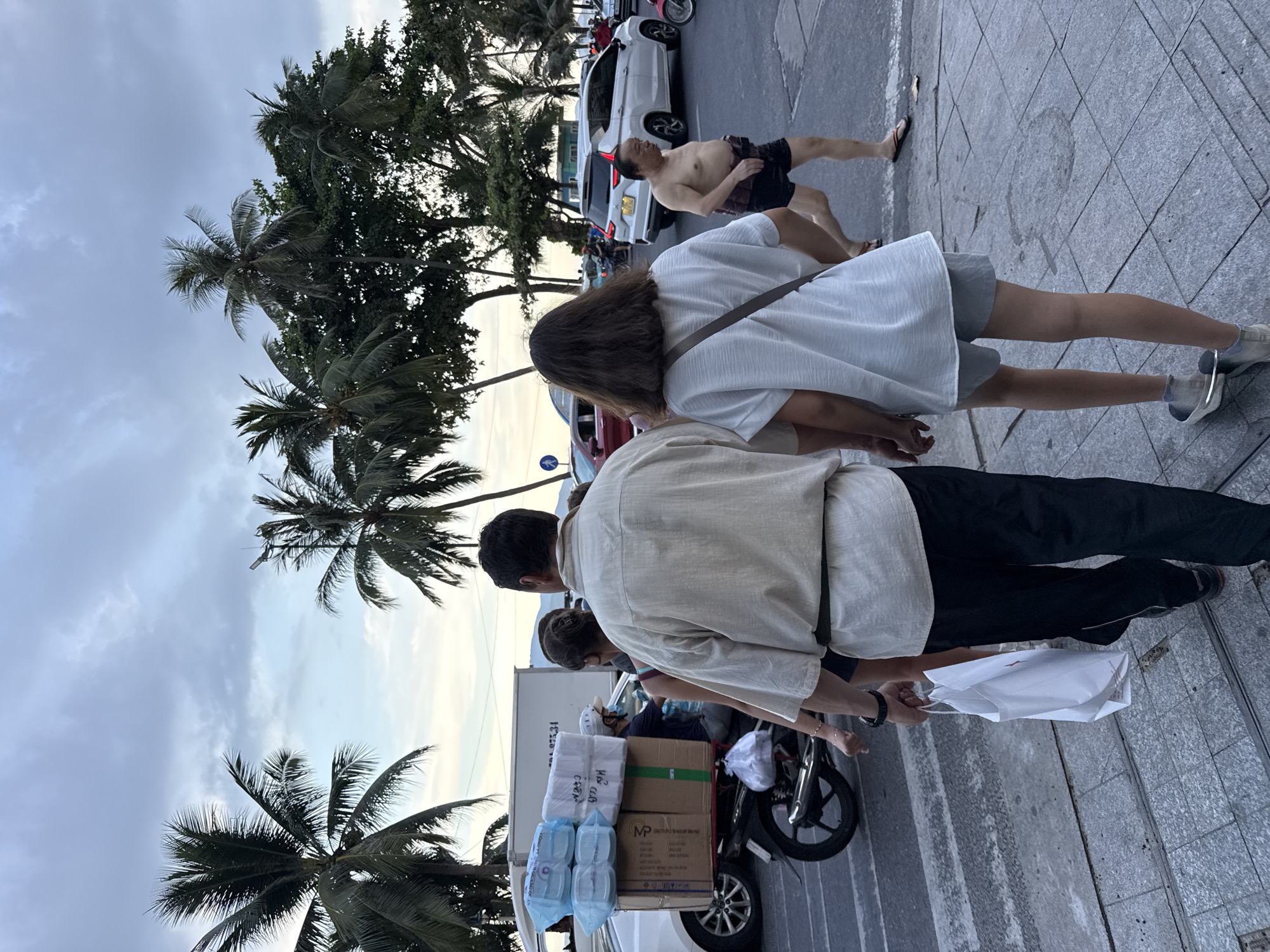 The image depicts a street scene in Nha Trang Ward, Vietnam. In the foreground, a man wearing a light-colored long-sleeved shirt and black pants walks with a white paper bag in his left hand. He is holding hands with a young woman in a white t-shirt and gray shorts. Behind them, to the left, a motorcycle is loaded with several boxes and blue packages. The motorcycle is driven by a person wearing a face mask and a white helmet. In the background, several cars, including a white SUV, are visible on the street, suggesting an urban environment. Palm trees are scattered throughout the scene, with a cloudy sky overhead. A shirtless man in black shorts is seen walking in the middle ground to the right.