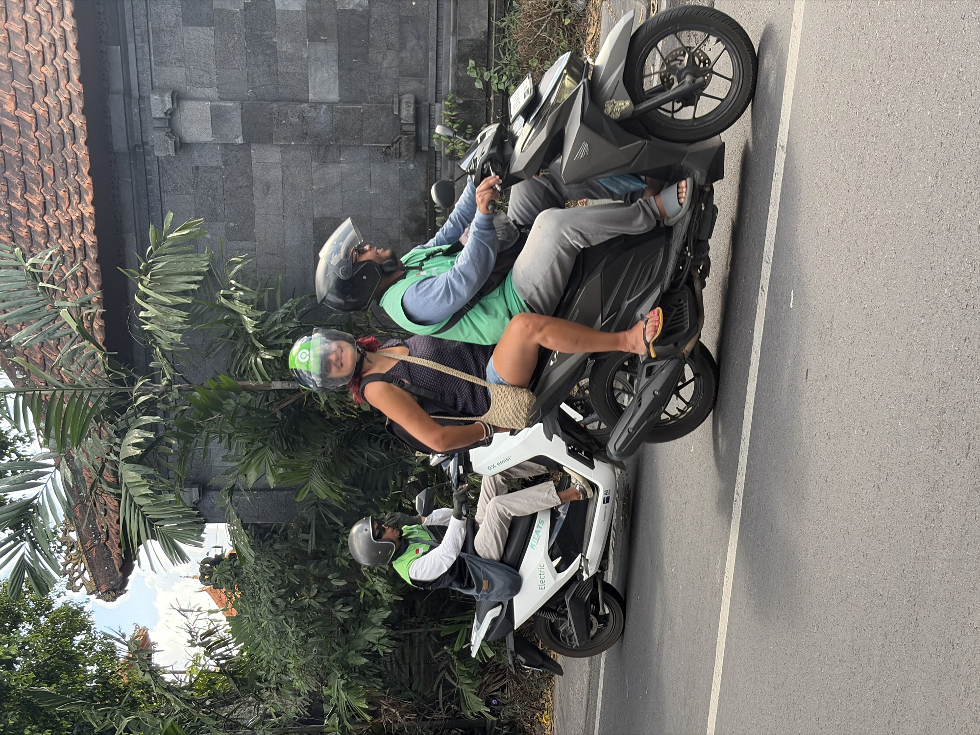 Two motorbikes are riding on a street in North Kuta, Indonesia. The first motorbike, a white electric scooter, is ridden by a man wearing a white long-sleeved shirt, a dark vest, beige pants, and a grey helmet. He has a gray bag slung over his shoulder and is wearing sunglasses. He is looking to his right. Behind him on the same scooter is a woman wearing a dark tank top and blue shorts, with a beige woven bag on her shoulder and a green helmet. She is looking forward. The second motorbike is a dark grey scooter. The man riding it is wearing a blue long-sleeved shirt and grey pants, with a black helmet and sunglasses. He is looking to his left. Behind him on the scooter is a woman wearing a dark top and jeans, with a green helmet. The motorbikes are on a paved road with a white line marking the lane. In the background, there is lush green foliage, a stone wall, and a red-tiled roof of a building. The sky is visible through the trees.