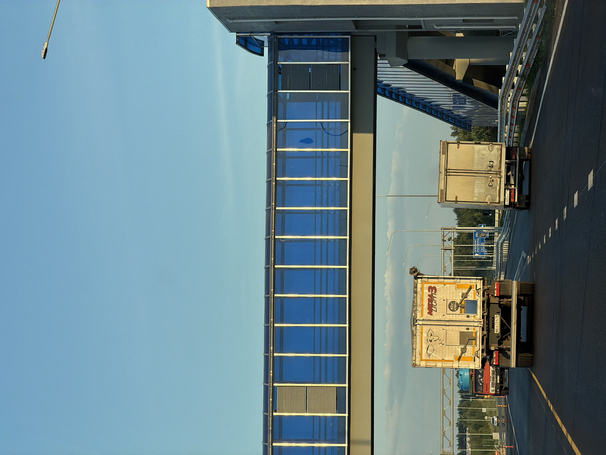 The image shows a highway overpass with a pedestrian walkway, seen from the perspective of a vehicle traveling behind two trucks. The walkway, with its blue glass panels and metal framing, spans across the highway. The sky is clear and blue, with some faint clouds visible in the distance. The highway is paved and has clear lane markings. The foreground shows the rear of two trucks, one white and one light-colored. The white truck has markings that read "MEGA 3 LIGHT" and a license plate that appears to be Russian. The environment is likely a semi-rural or suburban area, given the presence of trees lining the highway in the background. The overpass and highway suggest a well-developed transportation infrastructure. There are no people visible in the image. The scene appears to be captured during daylight, possibly in the late afternoon, due to the lighting and shadows.