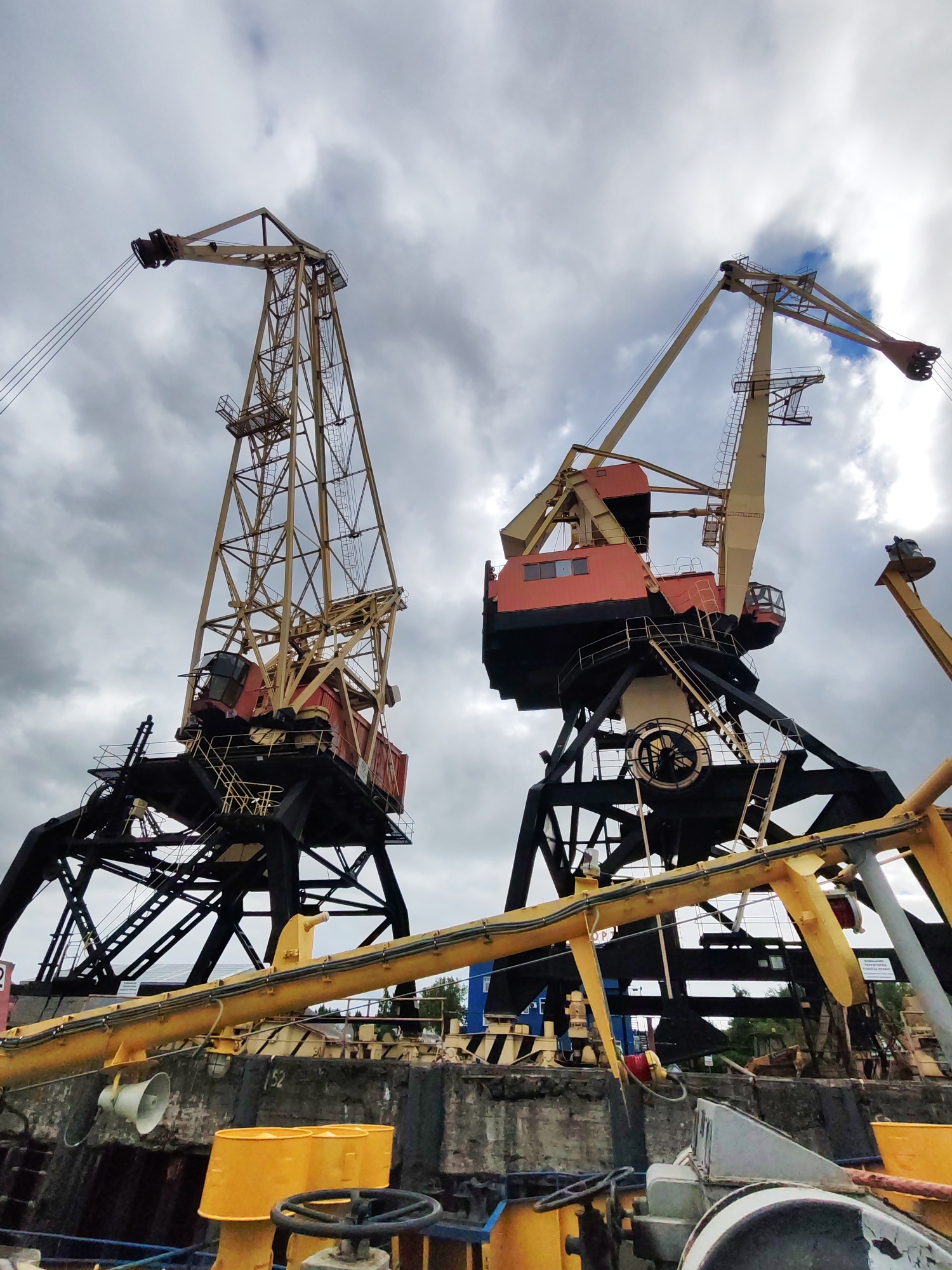 The image shows two large industrial cranes in a port, likely in the city of Rybinsk, Russia, based on the style of cranes common in former Soviet ports. Both cranes are painted with a combination of black, yellow, and red/orange colors. The crane on the left appears to have a more open lattice-style construction for its boom, while the crane on the right has a more solid, enclosed structure with a visible operator's cabin. The bases of the cranes are primarily black, made of heavy steel beams and girders. The sky above is overcast with heavy, gray clouds. Yellow piping is visible in the foreground and the concrete pier or dock is also visible. The overall impression is of a functional, but aging industrial port facility.