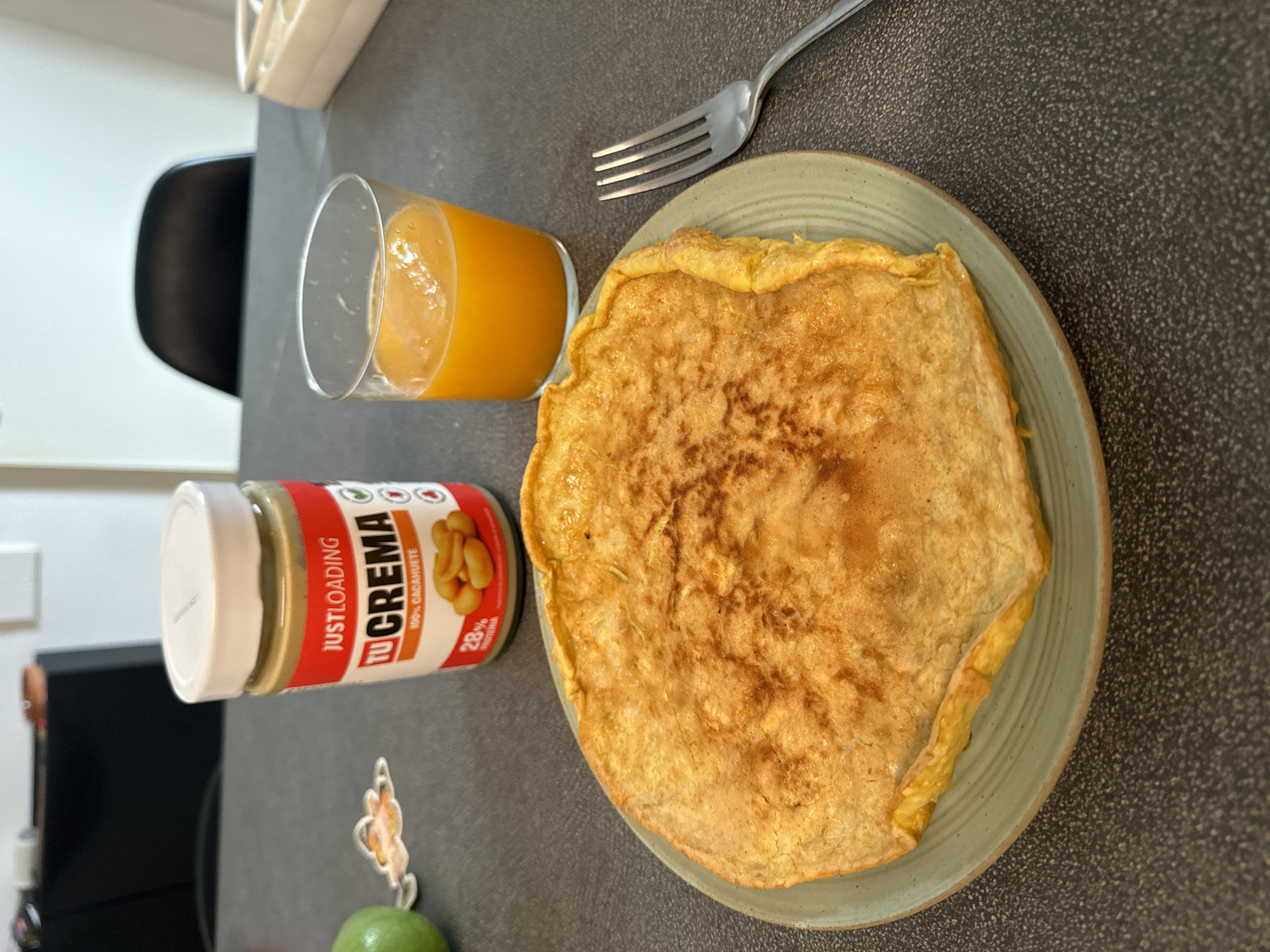 This media file displays a close-up, top-down view of a breakfast spread on a dark, speckled countertop. The scene is likely indoors, given the artificial lighting and the presence of kitchen items.
The primary subjects are an omelet, a glass of orange juice, and a jar of peanut butter. The omelet is golden brown and appears to be folded in half or possibly a crêpe, resting on a pale green, textured plate. A silver fork with four tines is positioned to the right of the omelet, suggesting it's ready to be eaten.
To the left of the omelet is a clear glass filled with vibrant orange juice. Beside the glass is a white jar with a red and white label. Visible text on the label includes "JUSTLOADING" in white letters on a red background, and "TU CREMA" in large, bold white letters. Below "TU CREMA," smaller text reads "100% CACAHUETE," and to the right, "28% PROTEINA." The label also features an image of peanuts.
Other objects in the background, partially visible, include a portion of a white shelf or cabinet and the back of a black chair. There is also a small, colorful sticker of a butterfly or fairy figure on the countertop. A small portion of a green fruit, possibly a lime or small apple, is visible at the bottom edge of the frame.
The overall impression is of a healthy and simple breakfast. The lighting is bright and casts minimal shadows, suggesting it could be daytime. The context of Murcia, Spain, might imply a warm climate, but this cannot be confirmed from the image alone. No people are visible. The setting appears to be a home kitchen or dining area.