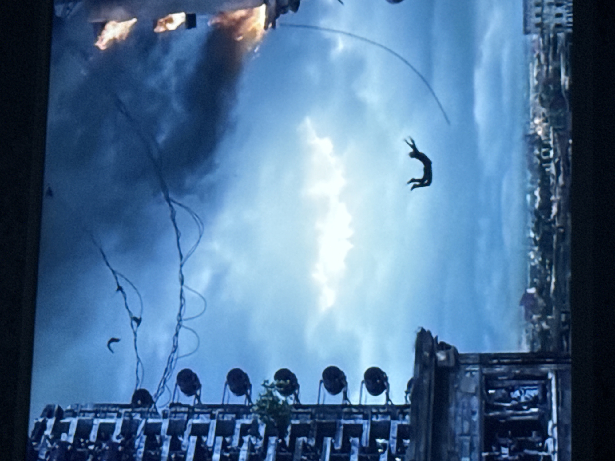 The image depicts a scene of destruction, potentially in an urban setting. A figure is falling mid-air against a backdrop of a cloudy sky. In the upper left corner, there is an explosion with fire and smoke. Below the figure, there's a building with what appear to be circular objects, possibly lights, lined up on the roof. There are wires or cables hanging in the air near the building. Buildings are seen in the background. It is not possible to confirm the location or city name from this image alone.