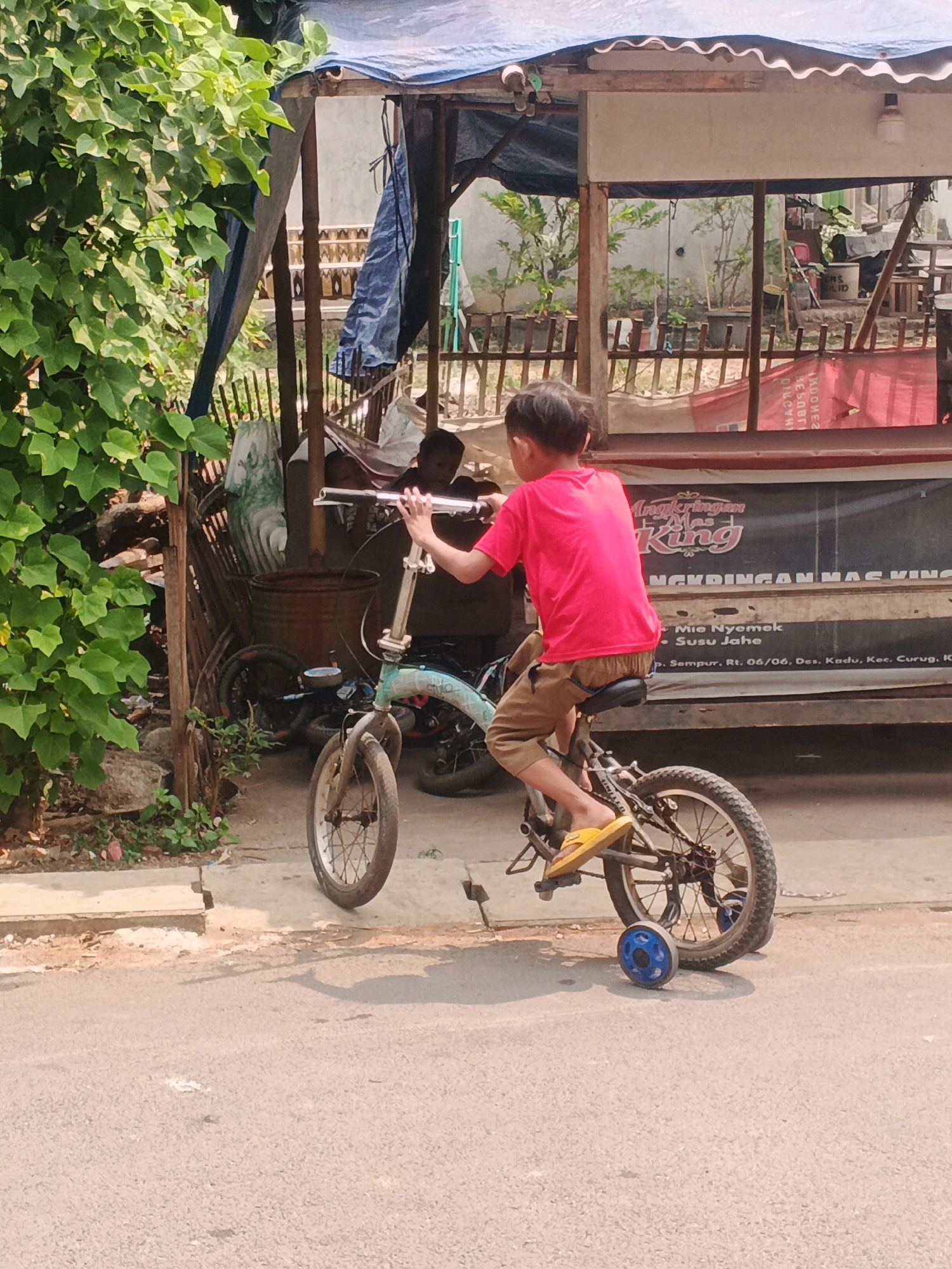 A young boy, wearing a bright pink t-shirt and tan shorts, rides a small, faded blue bicycle with training wheels on a paved street. He is in motion, with his hands on the handlebars and his feet on the pedals and footrests. The bicycle has a slightly worn appearance, with a blue training wheel attached to the rear wheel. Behind the boy, a rustic structure with a corrugated metal roof and blue tarpaulin sheeting is visible. This structure appears to be a roadside stall or workshop, with various items stored within, including large metal containers and wooden fencing. The surrounding environment suggests an outdoor setting, with lush green foliage encroaching on the scene from the left. The ground is a mix of asphalt and concrete. Text on a banner behind the boy, in Indonesian, reads "Angkringan Mas King" and provides an address: "Sempur, Rt. 06/06, Des. Kadu, Kec. Curug, K". This indicates the location is Kadu, Indonesia, likely in the Curug sub-district. The lighting suggests a sunny day.