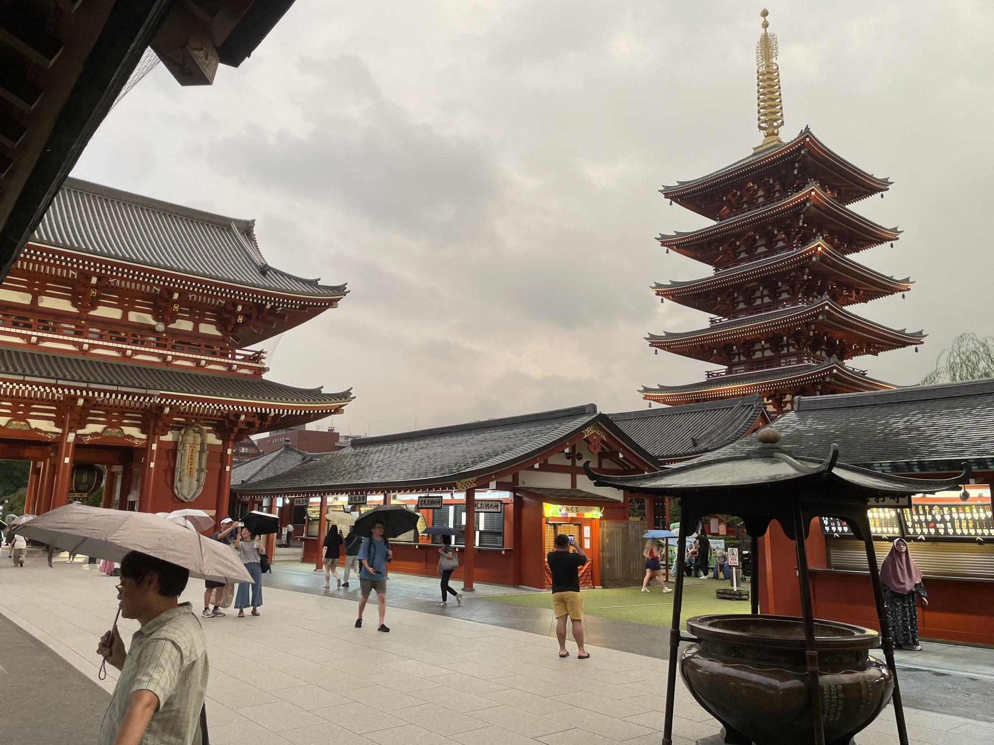 The image depicts the Senso-ji Temple in Taito, Japan, on an overcast day. The temple complex features traditional Japanese architecture, including red and white pagodas and ornate roofs. Several people are visible, many holding umbrellas, suggesting it might be raining or has recently rained. Tourists are walking around, some taking photos. One person in the foreground is holding a large umbrella over their head and is looking to the left. In the background, a five-story pagoda stands tall against the cloudy sky. The ground is paved with light-colored tiles. There are stalls or shops with signage, and a large bronze incense burner is in the foreground on the right. The overall atmosphere appears to be that of a bustling tourist site despite the weather.