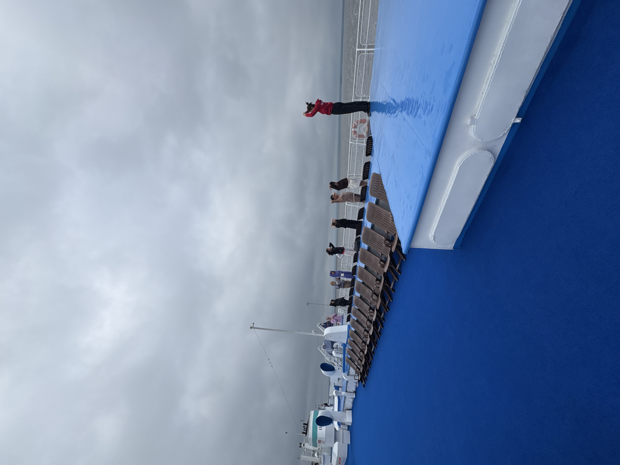 A group of people are on the deck of a ferry or cruise ship, with a vast expanse of cloudy sky overhead. The deck is covered in a vibrant blue material, and rows of empty brown lounge chairs are lined up. In the foreground, the blue deck slopes downwards and to the left. In the mid-ground, a person in a red jacket stands on the blue deck, facing away from the camera with their hands raised above their head. Several other individuals, dressed in casual clothing, are standing in a line on the deck behind the person in red, also with their hands raised. To the left, a portion of the ship's superstructure is visible, painted white with blue accents and green pipes. The horizon line, where the water meets the sky, is barely discernible due to the overcast conditions. The overall atmosphere is subdued, possibly due to the cloudy weather. This scene is not specifically identifiable as being in the city of Laishevo, Russia, as there are no distinguishing landmarks or contextual clues present in the image to confirm its location.