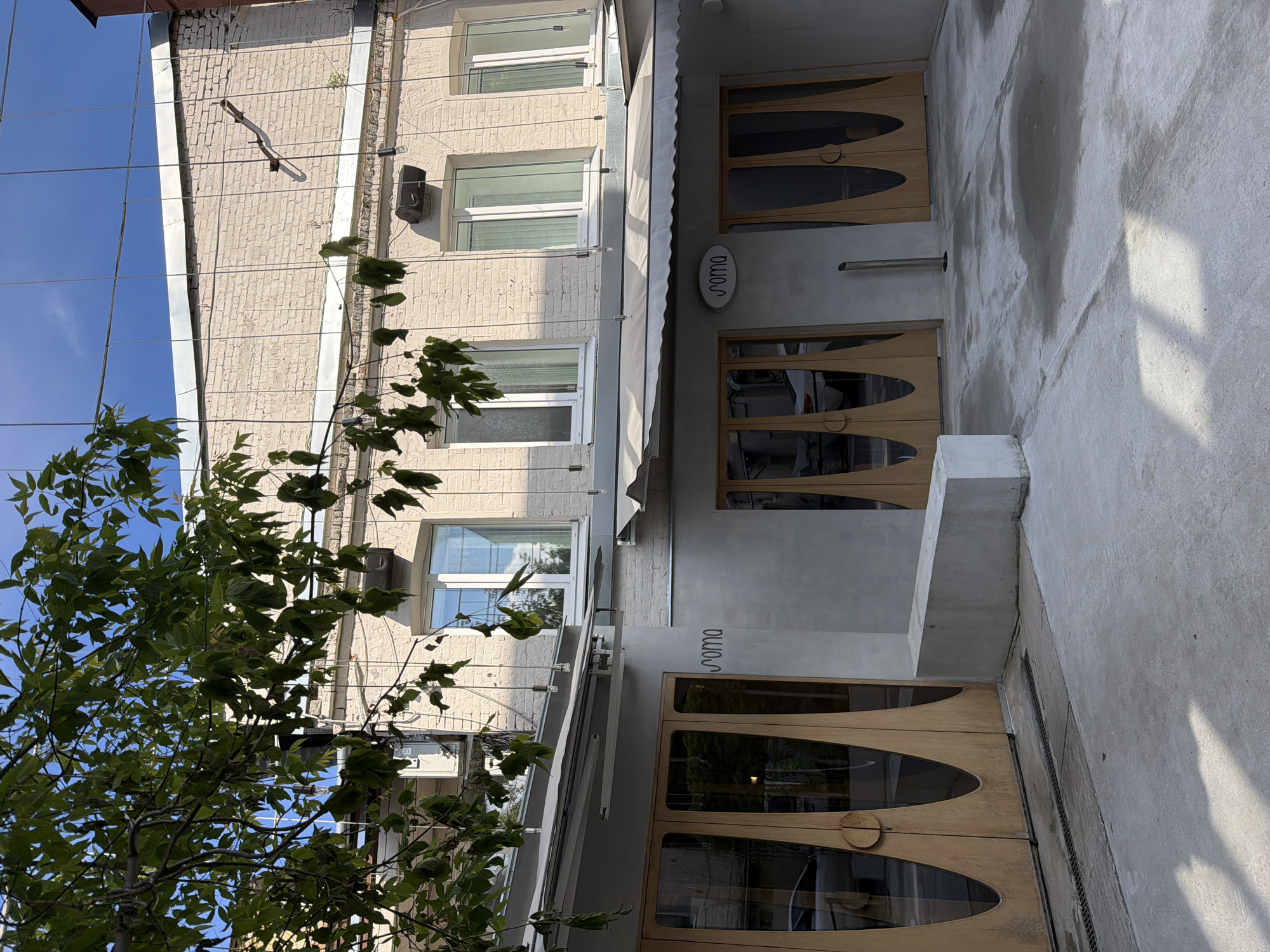 The image shows a building facade with three floors and a street-level entrance. The building is constructed of light-colored brick. The windows are rectangular with white frames. A tree with green leaves partially obscures the view of the building. The entrance has three sets of wooden double doors, each with a large, oval glass panel. Above each entrance is a gray sign that reads "roma" in lowercase. A gray awning extends over the entrance. The street is made of gray concrete. The building is located in Moscow, Russia.