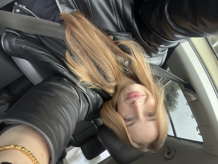 A female individual is depicted taking a selfie inside a vehicle. She has long, light brown hair, light-colored eyes, and full, glossy lips, exhibiting a slight smile. She is wearing a black leather jacket over a dark garment, with a dark-colored seatbelt secured diagonally across her chest. A gold-colored, multi-link bracelet is visible on her right wrist. The vehicle interior features dark, likely leather, upholstery on the front and rear passenger seats. Through the car window, an exterior environment shows trees and an overcast sky. No specific geographical landmarks or features are discernible within the image to confirm the stated location of Medinski Krš, Montenegro.