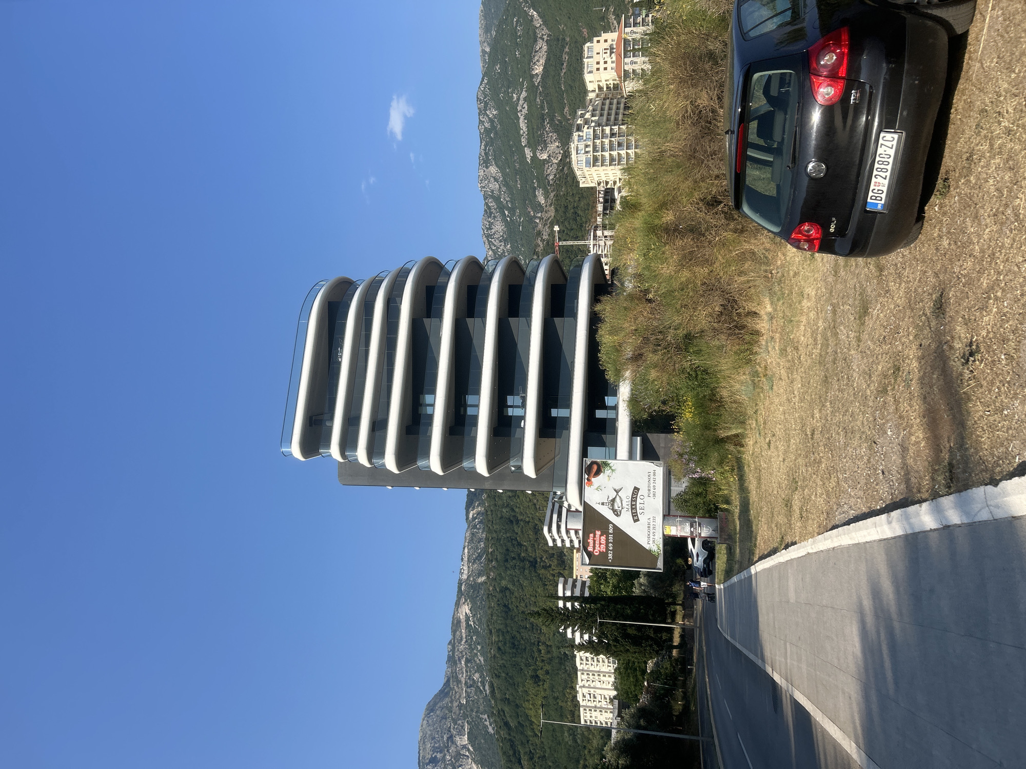 This media file captures a sunny day in Boreti, Montenegro, with the distinctive architecture of a modern building dominating the left side of the frame. The building features a curved, layered facade with white horizontal elements contrasting with dark glass sections. In the background, lush green mountains rise dramatically, dotted with buildings, suggesting a hillside community. A black Volkswagen Golf is parked on the right side of a paved road, its license plate clearly visible as BG 2880-ZC. The ground is covered in dry, yellowish grass, indicative of a warm, dry climate.
A billboard near the modern building advertises "MALO RIBARSKO SELO" with contact numbers and mentions "Budva Opening" on September 29th, suggesting a business opening event. The sky is a clear, vibrant blue with a few wispy clouds. The scene appears to be set during the daytime, likely late morning or early afternoon, given the bright sunlight and shadows. The overall impression is one of a scenic coastal location with modern development set against a natural, mountainous backdrop.