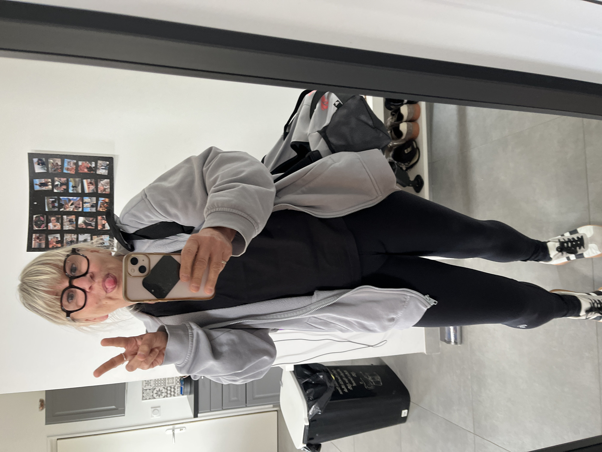 A person, likely female, is taking a selfie in a mirror. They are wearing glasses, a grey hoodie, a black top, and black leggings, and sporting white and black sneakers. They are making a peace sign with one hand and sticking their tongue out while holding a smartphone. The reflection reveals a kitchen or kitchenette area with grey cabinets and white doors. A photo collage is visible on the wall behind them. A trash can is also visible near the base of the mirror. The city of Chambœuf, France is not discernible from the visual information.
