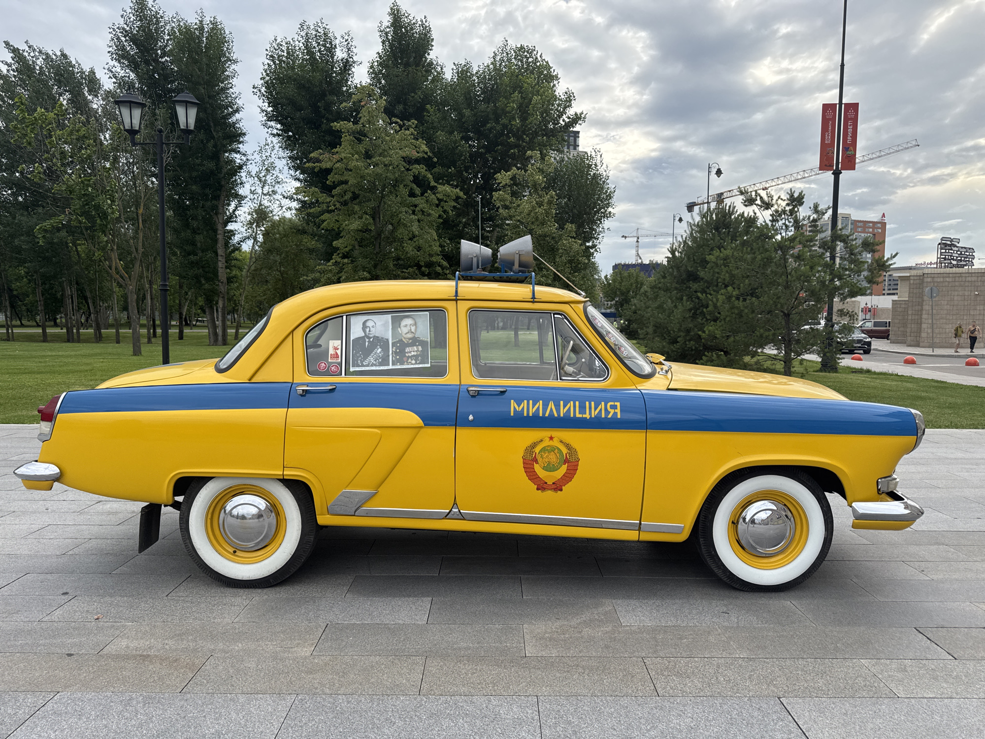 This is a photograph of a vintage yellow and blue police car, likely a GAZ-24 "Volga," parked on a paved walkway in a city park. The car has "МИЛИЦИЯ" (Militiya) written in Cyrillic on its side, along with a red and gold emblem. Two framed portraits of men in military uniforms are visible through the passenger-side window. A pair of bullhorn-like speakers are mounted on the roof of the car. In the background, there are mature trees, a lamppost with two lights, and in the distance, several buildings, including a tall red and white structure, and construction cranes. The sky is overcast. The location appears to be in a park or public space, possibly in Moscow, Russia, given the signage and architectural style. There are no people in the immediate vicinity of the car.