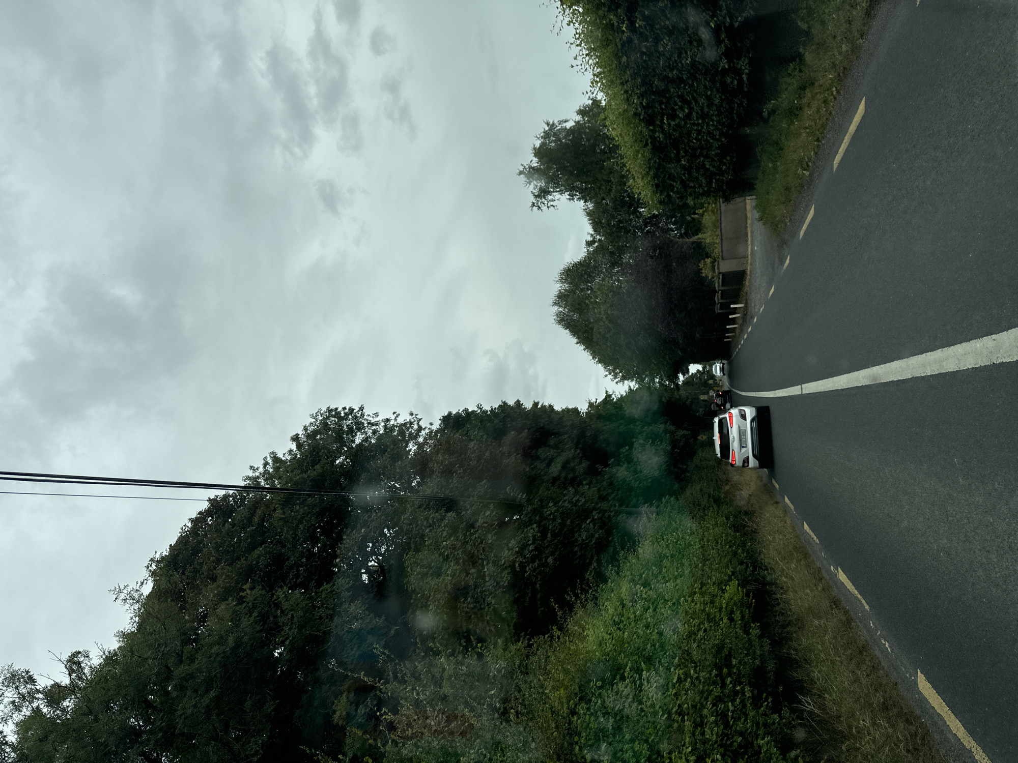 The image captures a view from a vehicle traveling on a rural road. The road is paved and marked with a white line down the center and yellow lines on the right edge. Dense green foliage and trees line both sides of the road, creating a natural tunnel effect. A white car is visible ahead on the right side of the road, followed by another vehicle further in the distance. The sky is overcast and appears to be cloudy. The scene suggests a somewhat overcast day in a rural setting. The location is described as being in Athgarvan, Ireland.