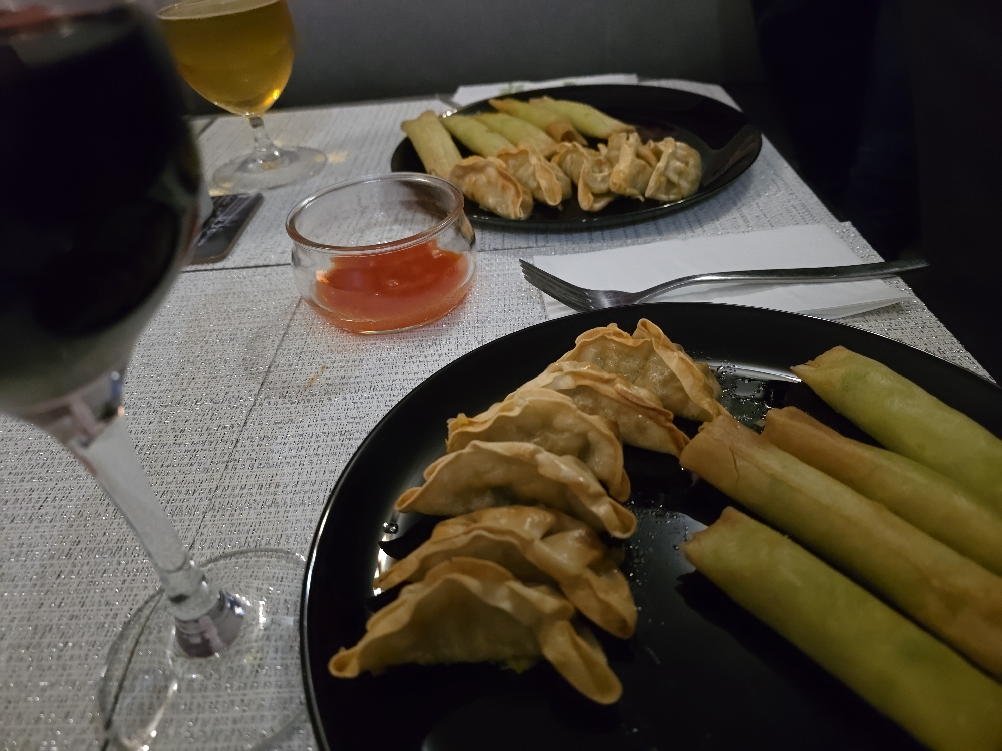 The image shows a table setting with two black plates of food, a small dish of red sauce, two glasses of alcoholic beverages, and a fork.
On the lower plate, there are several fried dumplings arranged along one side and a set of fried spring rolls along the other. The upper plate also contains both fried dumplings and spring rolls. A small glass bowl filled with a red sauce is positioned near the upper plate.
One glass contains a dark red liquid (likely wine), while the other holds a light yellow liquid (possibly beer or white wine). A fork rests on a white napkin near the upper plate. The table has a textured white surface. The backdrop is indistinct but seems to indicate an indoor setting, with dark elements suggesting furniture or a wall.
The location cannot be ascertained from this image. There is nothing to indicate that this image was taken in Varna, Bulgaria.