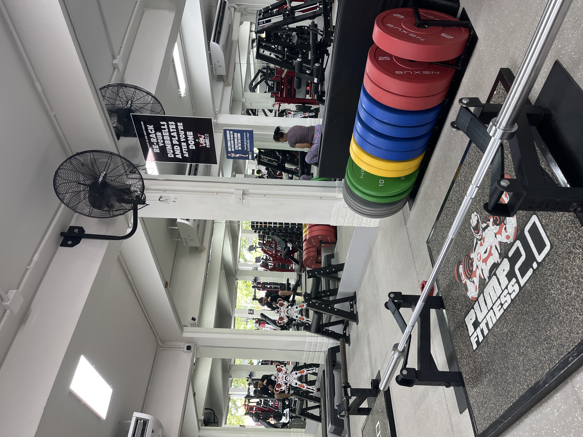 This is a wide-angle, slightly tilted, eye-level shot of the interior of a brightly lit gym in Chiang Mai, Thailand. The scene is dominated by exercise equipment, including weight racks, barbells, and a large stack of colorful weight plates. Two black oscillating fans are mounted on the wall, likely to keep the space cool.
In the background, several people are engaged in workouts, with one woman in the middle ground sitting on a bench and others visible on gym equipment reflected in the mirrors lining the walls. The overall atmosphere appears to be one of focused activity.
Prominent signage is visible, including a black and white sign with bold text that reads "RE-RACK YOUR DUMBBELLS AND PLATES AFTER YOU'RE DONE" and another sign in both English and Thai that mentions female members are welcome to use the stretching area on the 5th floor. A logo for "PUMP FITNESS 2.0" is displayed on a mat in the foreground. The clean, modern aesthetic of the gym, combined with the presence of multiple types of weightlifting equipment, suggests a well-equipped facility. The time of day is likely daytime, given the bright lighting and the appearance of natural light filtering in from windows or open spaces.