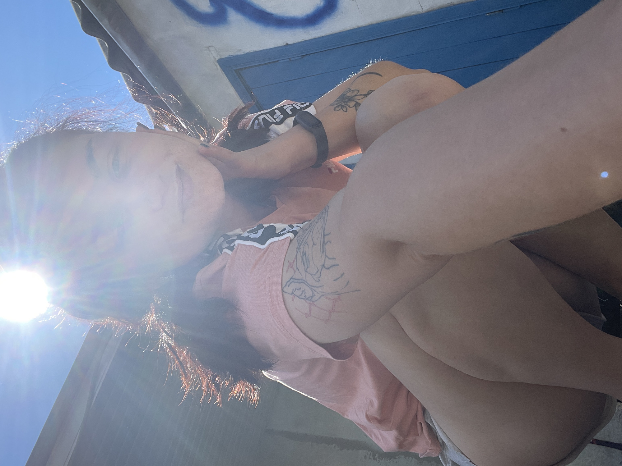 A low-angle, close-up selfie shows a young woman with dark hair posing outdoors. She is wearing a pink t-shirt and has tattoos on her left arm and thigh. The sun is shining brightly, creating a lens flare that partially obscures her face. Her hair is catching the light and appears to have copper highlights.
Behind her, a blue wooden structure with some graffiti is visible. The sky is clear and blue. The overall impression is of a sunny day, possibly during the afternoon given the angle of the sun. The woman appears relaxed and is looking at the camera. The location context provided is Dnipro, Ukraine.