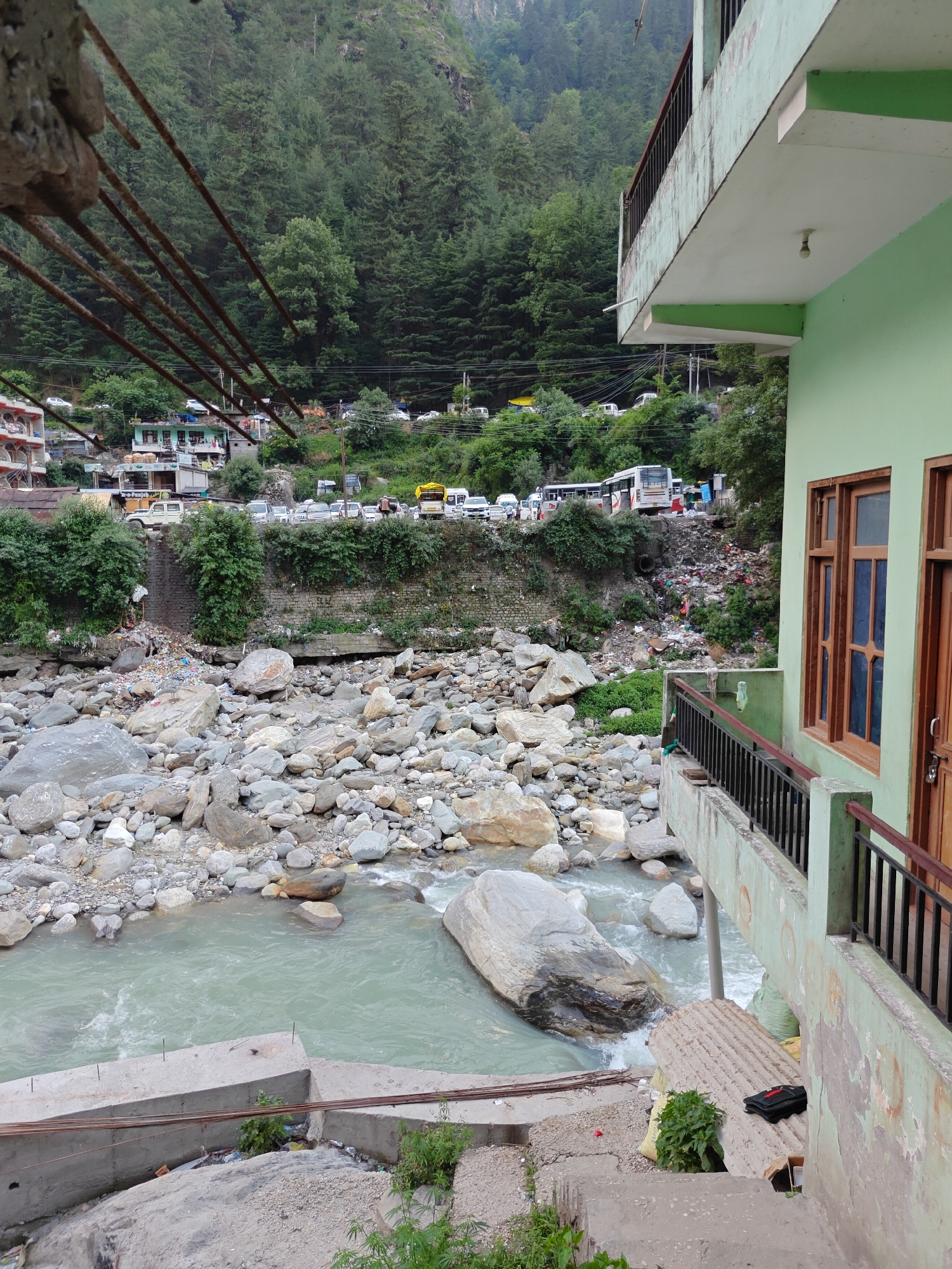 This image depicts a mountainous landscape in Manikaran, India, characterized by a river, dense forests, and human settlements. The perspective is from an elevated position, looking down towards a river and across to an opposing hillside.
In the foreground, on the right, part of a light green building is visible, featuring wooden-framed windows and a concrete balcony with a black metal railing. A single light bulb hangs from the underside of the balcony. In the lower left, several rusty rebar rods protrude from a concrete structure, suggesting an unfinished construction or infrastructure.
The middle ground is dominated by a river with light blue-green water, flowing over and around numerous large and small grey boulders and rocks. The water appears swift in places, creating small rapids. The river banks below the road show a concerning accumulation of trash and debris, including plastic bags and various refuse.
Across the river, a winding road is bustling with activity, packed with a variety of vehicles including cars, buses, and what appears to be a white jeep with "A-O-PUNJAB" clearly visible on its side. Several multi-story buildings and other structures are built into the steep hillside alongside the road. Behind these structures, the mountain rises sharply, densely covered with dark green evergreen trees, forming a thick forest.
The weather appears to be overcast, with no direct sunlight casting harsh shadows. The overall scene captures a vibrant yet somewhat cluttered mountain town, where natural beauty coexists with significant human impact and infrastructure.
