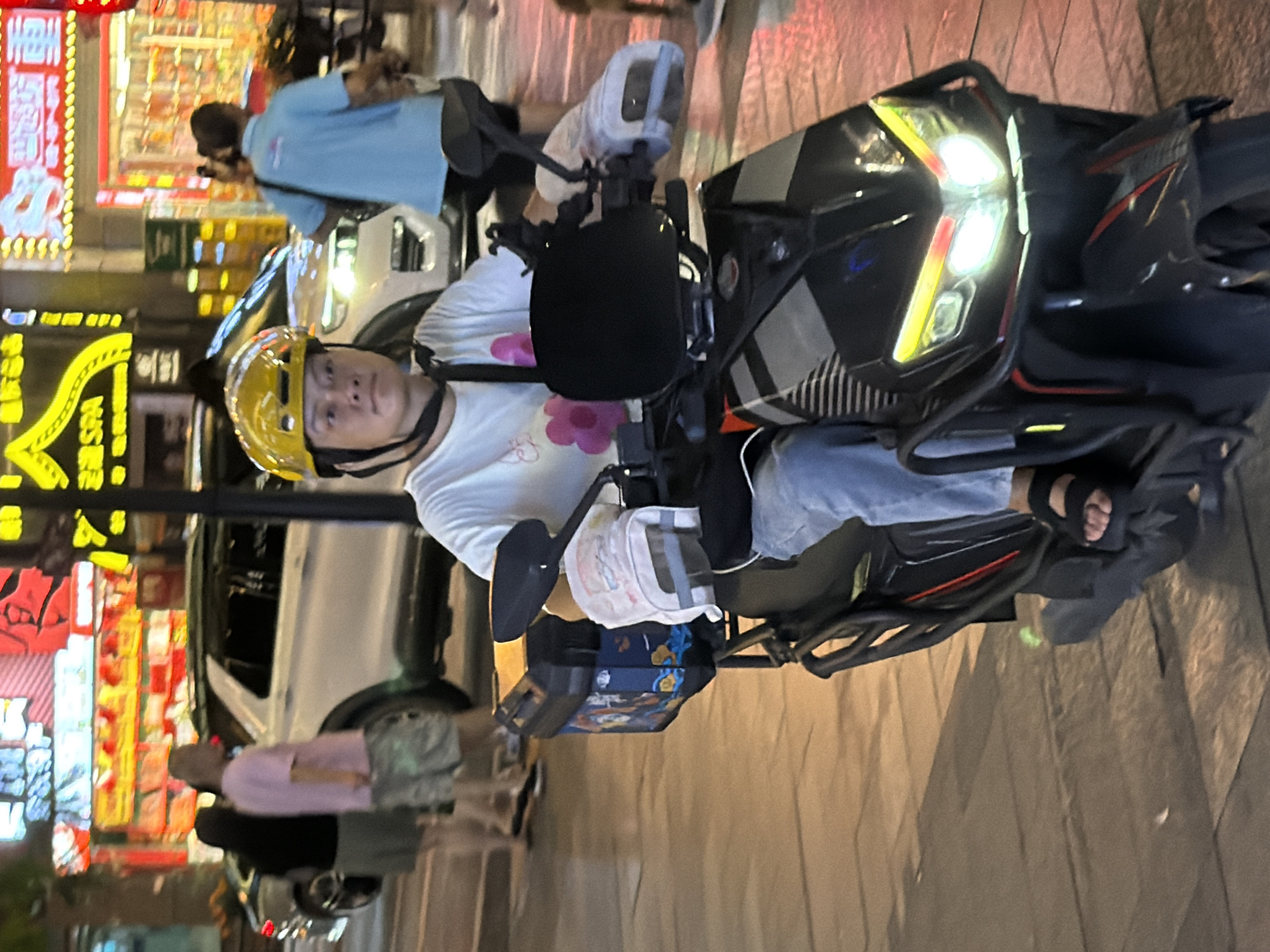 In Yuzhong District, China, a person wearing a yellow helmet and a white t-shirt with pink floral designs is riding a black electric scooter. The scooter has a black storage box on the back adorned with a colorful graphic. The rider is wearing blue jeans and black sandals. The scooter's headlights are illuminated, casting a bright light on the pavement. In the background, illuminated signs and a white SUV are visible, suggesting an urban street environment at night.
