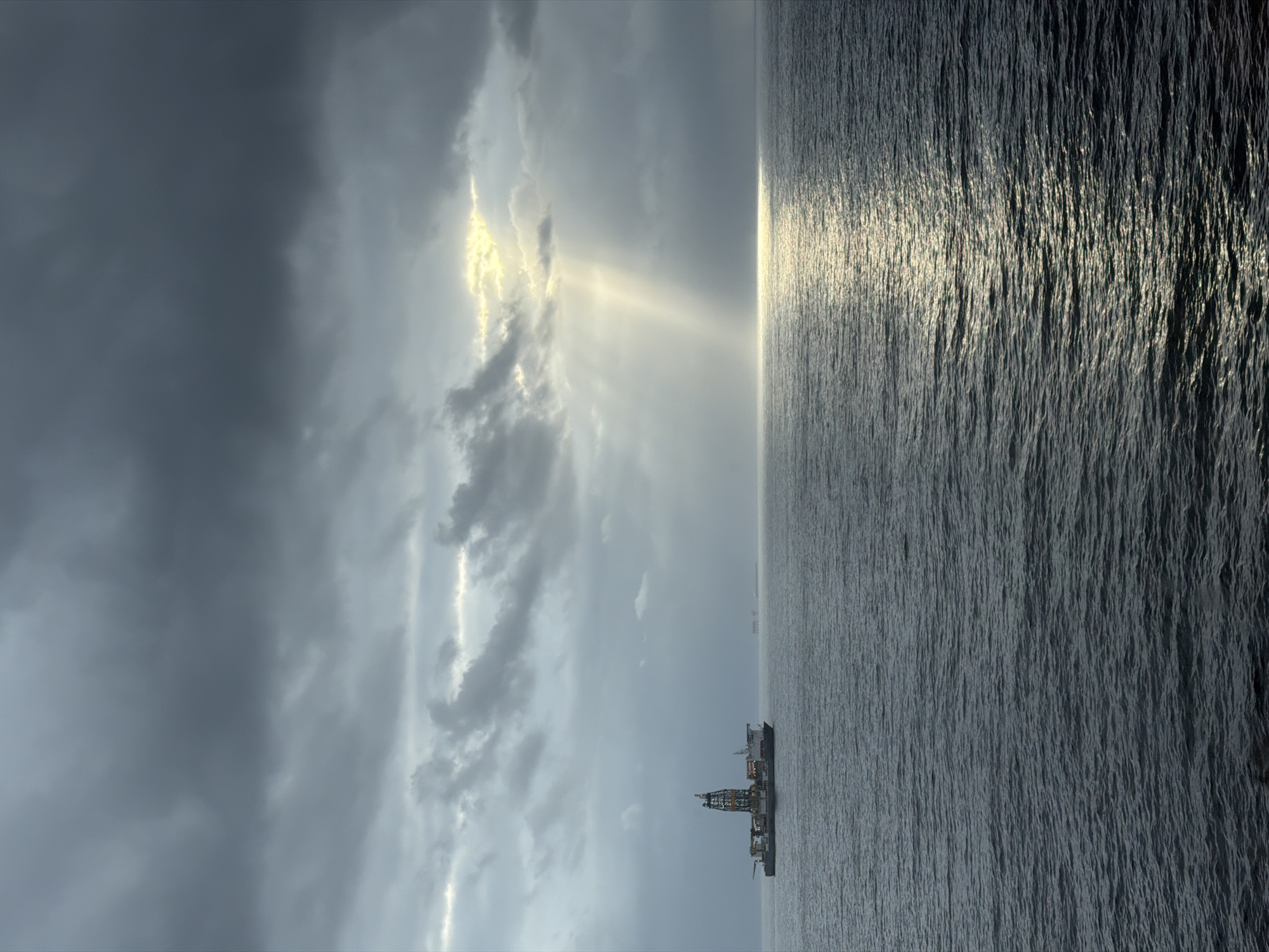 A dramatic seascape unfolds under a vast, cloudy sky, with the horizon line dividing the scene horizontally. The upper half is dominated by an expansive sky filled with varied clouds; dark, heavy formations occupy the left side, transitioning to lighter, more broken clouds towards the right. Striking crepuscular rays of sunlight pierce through gaps in the clouds, creating prominent beams of light that stretch downwards and illuminate sections of the cloud cover. A particularly bright, golden band of light is visible higher within the cloud layer.
The lower half of the image depicts a wide expanse of ocean, its surface textured by countless ripples and small waves. A shimmering path of sunlight extends across the water from the horizon towards the viewer, reflecting the bright light from the sky. On the distant horizon, a large, industrial-looking vessel, possibly an offshore support ship or drilling rig, is visible, featuring a tall, distinctive structure in its center. The lighting suggests either early morning or late afternoon, due to the dramatic sunrays and the bright reflection on the water. The overall scene conveys a sense of natural grandeur and vastness over the open sea. There is no visible text in the image.