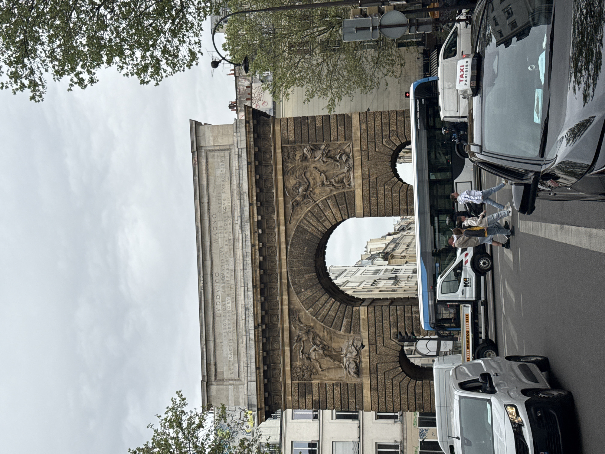 The image shows the Porte Saint-Martin in Paris, France. It is a stone archway with carvings and inscriptions. People are crossing the street in front of it. A white van and a black car are parked nearby, along with a Taxi Parisien vehicle. Trees are seen on either side of the archway. The sky is overcast.