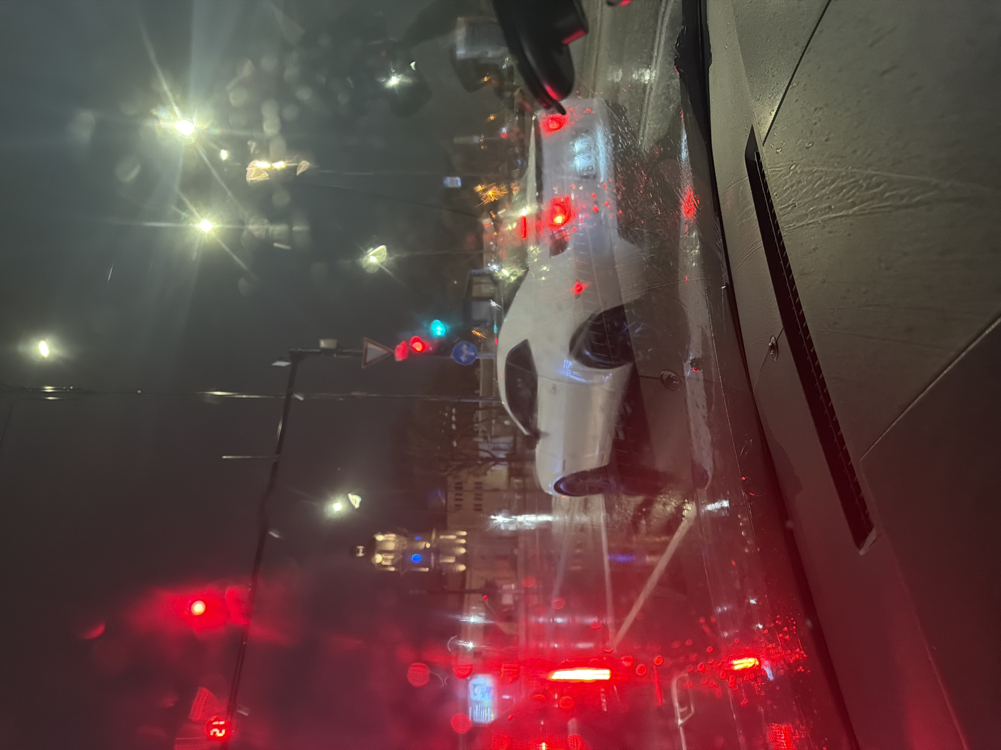 The image shows a nighttime view from inside a vehicle, looking out through a rain-streaked windshield. The foreground is dominated by the interior of the car. Through the windshield, a wet street scene is visible, illuminated by streetlights, traffic lights, and the headlights of other vehicles. A white car is partially visible to the right, appearing to be adjacent to the vehicle from which the photo was taken. In the background, there are buildings, some of which are brightly lit. The presence of traffic lights suggests an urban setting, likely a street in Varna, Bulgaria. The red lights of other vehicles are prominent in the lower part of the frame.
